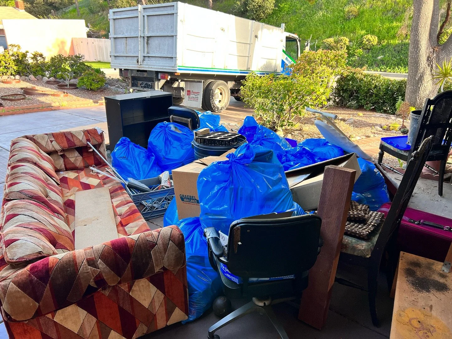 An outdoor view of a cluttered porch with various items including a colorful upholstered couch, black office chair, black chair, red suitcase, wooden piece, and many blue trash bags filled with items. In the background, a white truck is parked on the street, with greenery and trees around.