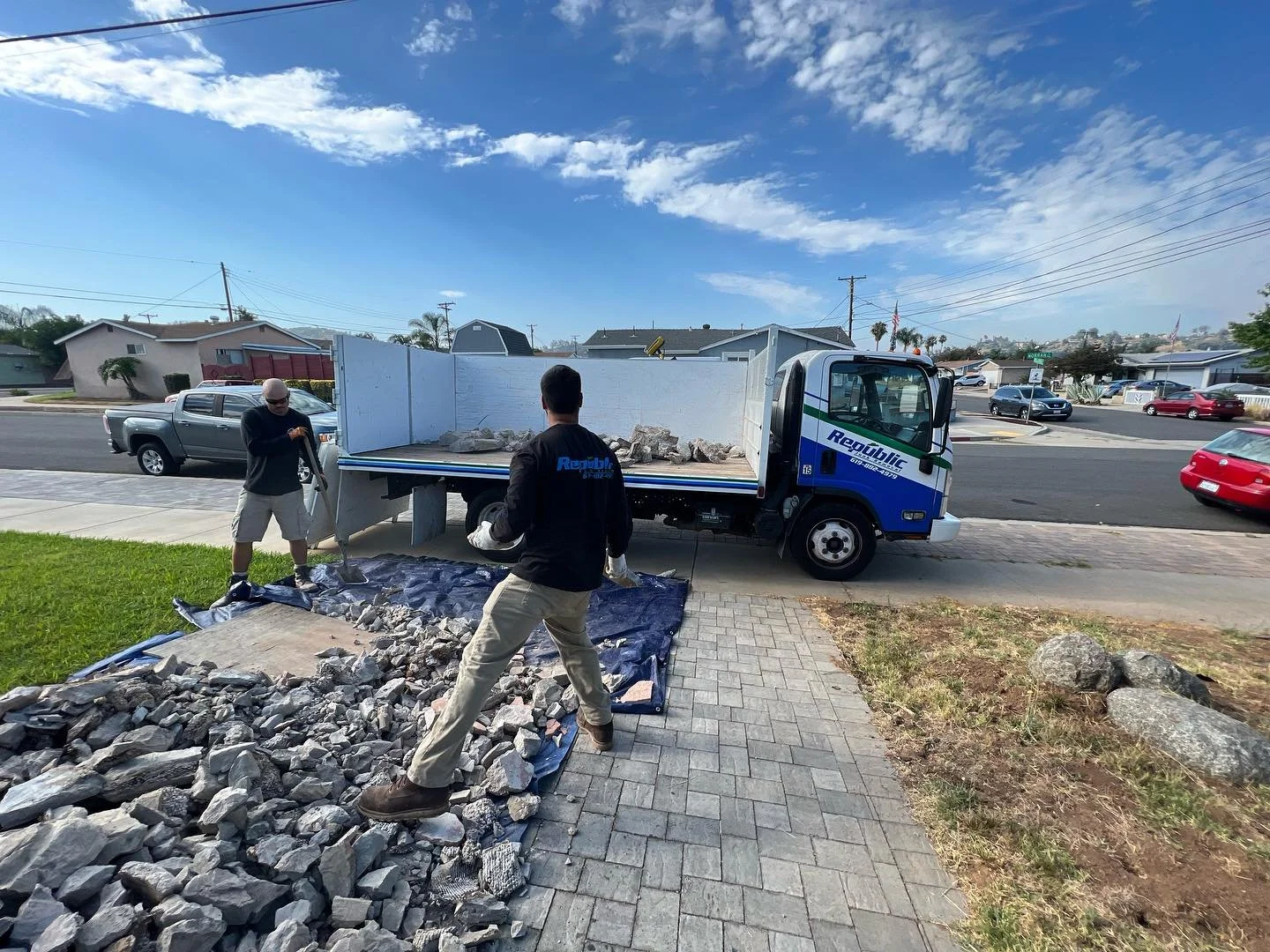 Two workers unloading rocks from a roadside truck onto a tarp on a sidewalk in a residential area under a blue sky with white clouds.