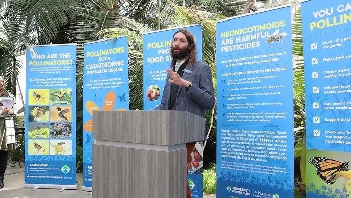 Man speaking at a podium outdoors, surrounded by informational posters about pollinators and pesticides, with palm trees in the background.