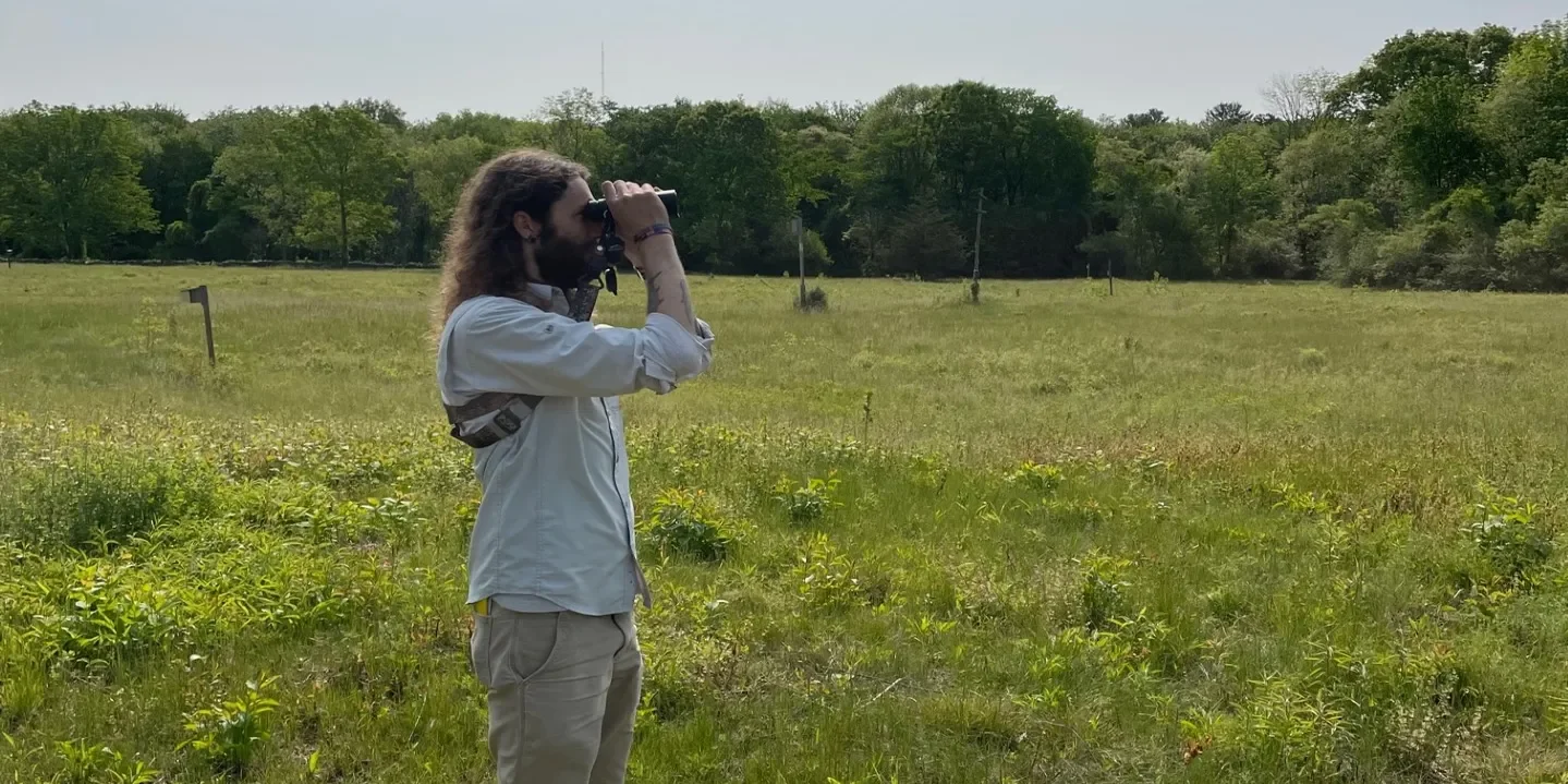 A person with long curly hair and a beard looking through binoculars in an open grassy field with trees in the background.