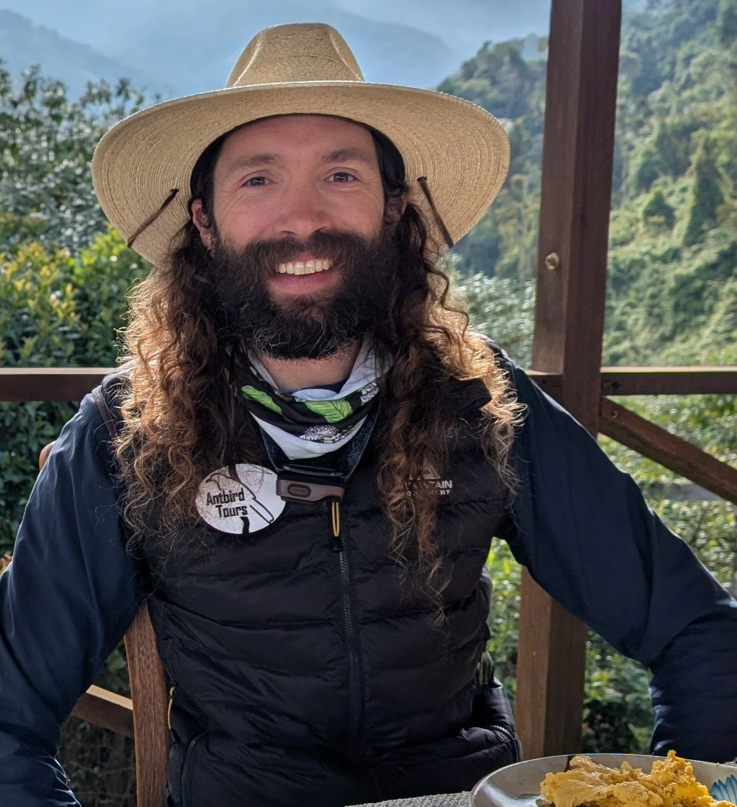 A man with long curly hair and a beard wearing a wide-brimmed straw hat, outdoor setting with green mountains in the background, sitting at a table with scrambled eggs.