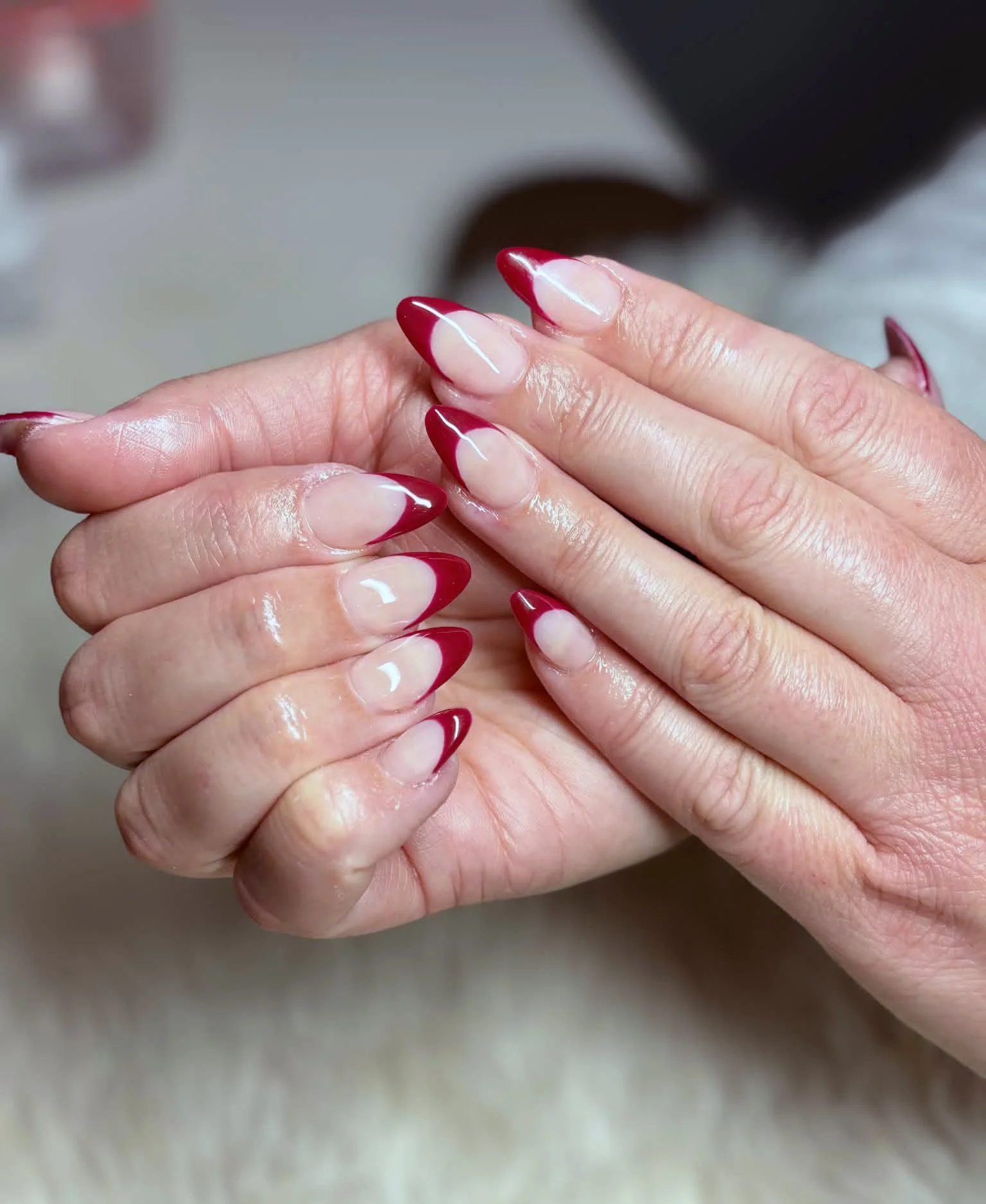 Close-up of hands with well-manicured nails featuring red French tips, with glossy finish, resting on a soft surface.