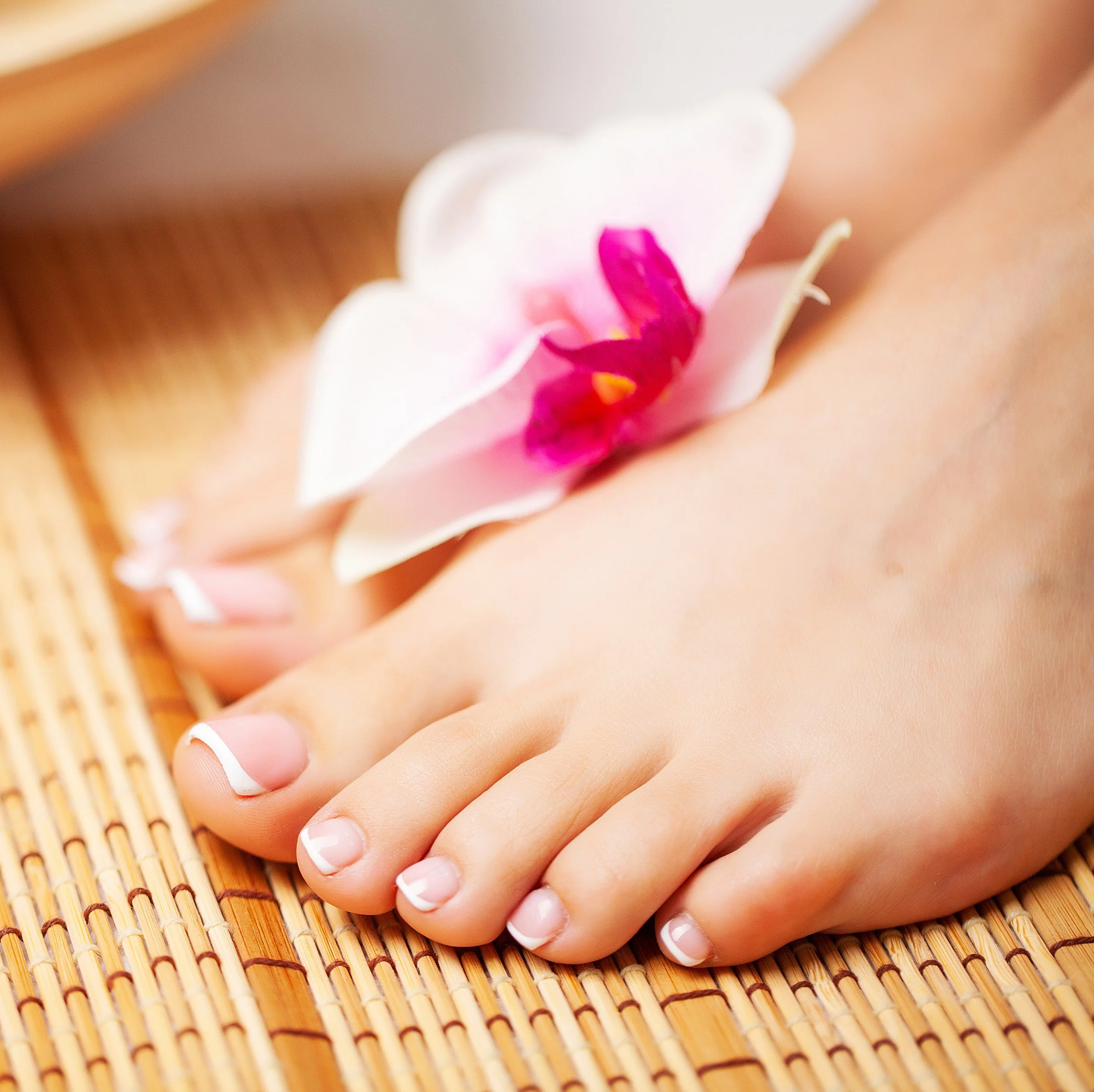 Close-up of a person's toes with French pedicure, on a bamboo mat, with a white orchid flower resting on the toes.