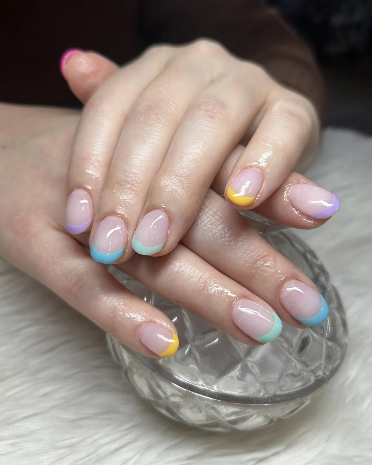 Close-up of hands with pastel-colored French manicure nails resting on a glass dish with ice, on a white fuzzy surface.