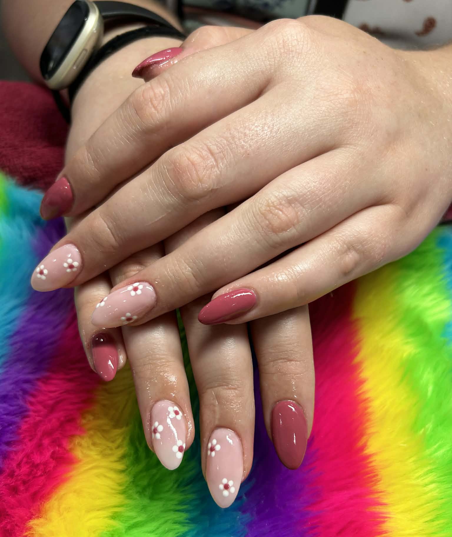 Hands with manicured nails resting on a rainbow-colored furry fabric. The nails are painted in shades of pink, with some nails featuring white floral designs.