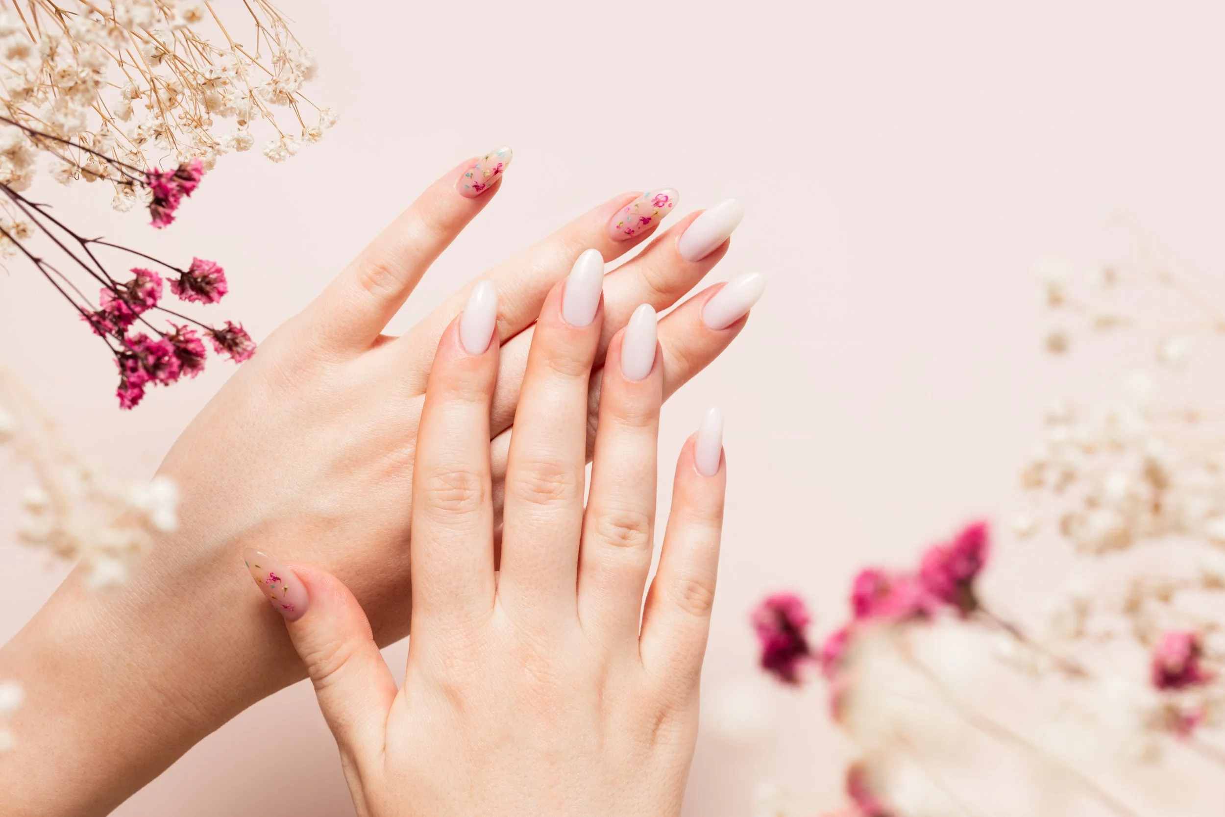 Close-up of a woman’s hands with manicured nails, one hand resting on top of the other, surrounded by small pink and white flowers.
