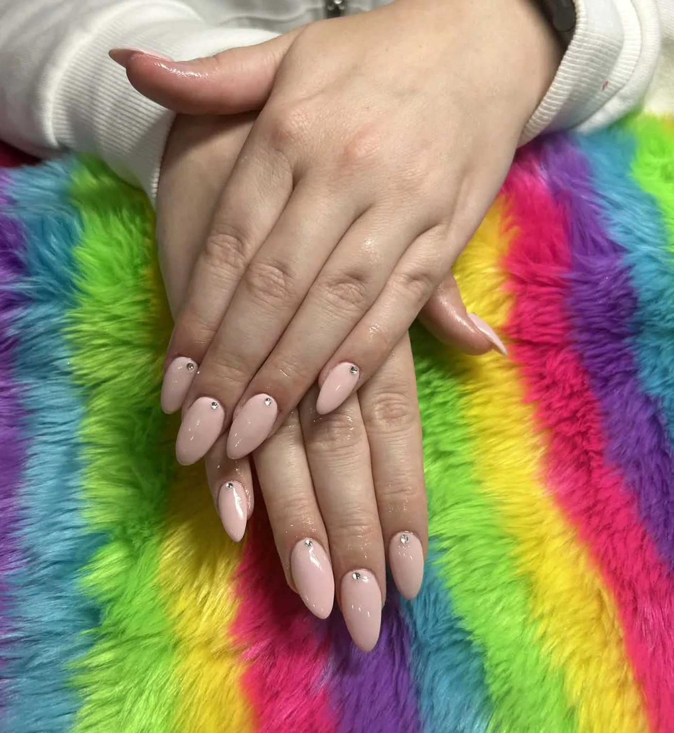 Close-up of a person's hands with manicured nails and small rhinestones, resting on a vibrant rainbow-colored furry blanket.