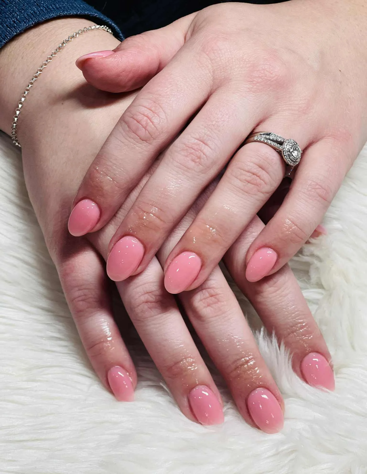 Close-up of two hands with pink manicured nails, wearing a diamond ring and a silver bracelet, resting on a white furry surface.