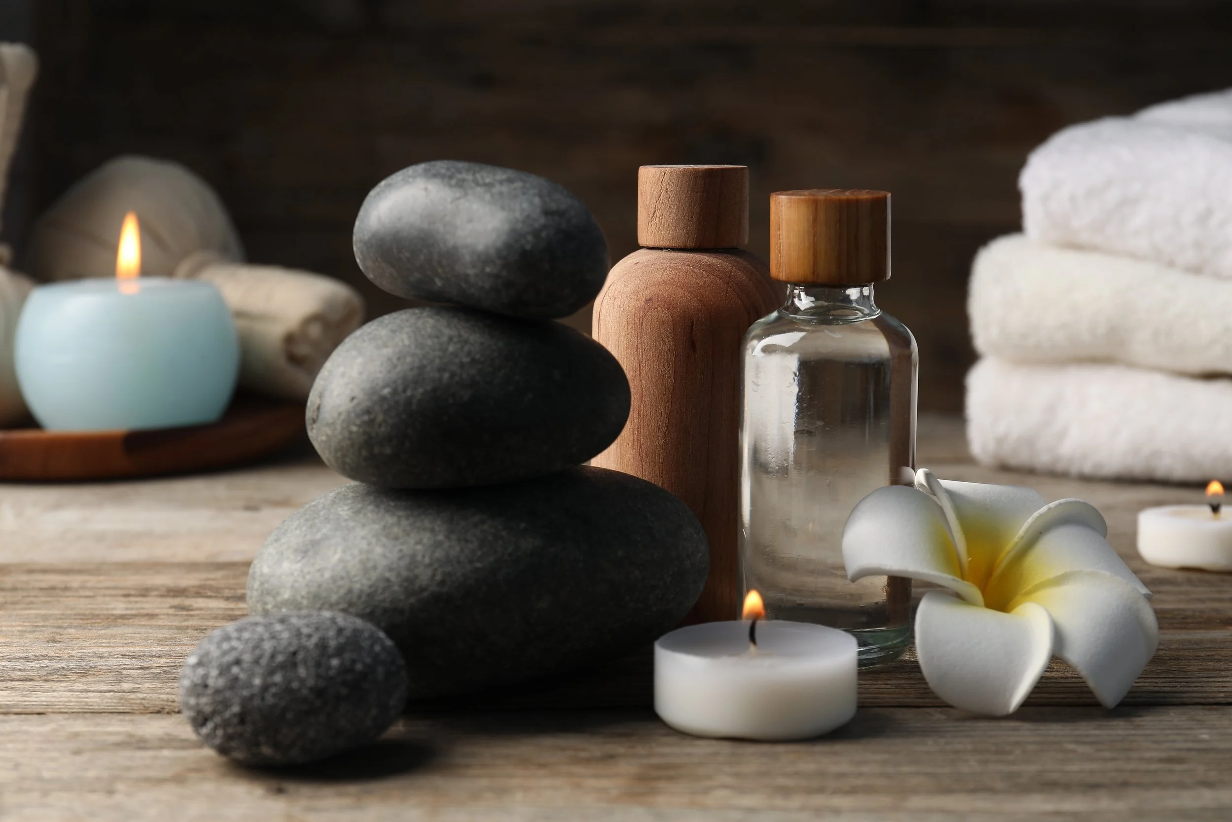 Spa setting with balanced gray stones, wooden bottles, a glass vial, white towels, candles, and a white flower.