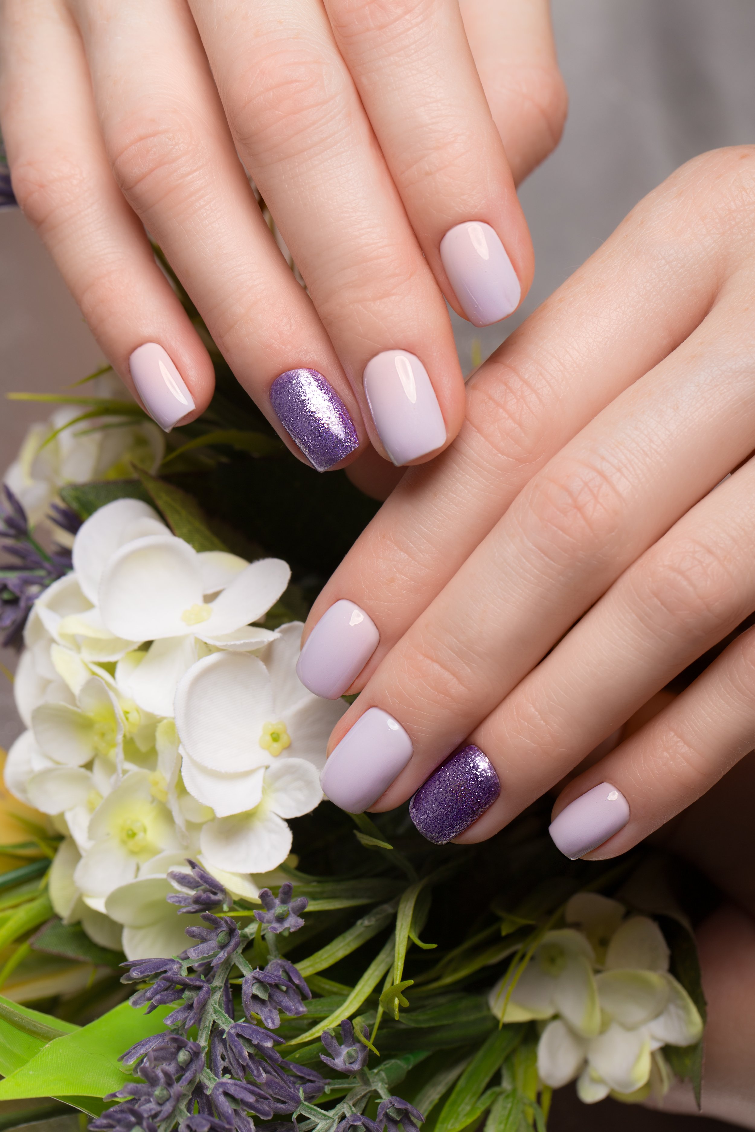 Close-up of manicured hands with white and purple glitter nail polish, surrounded by white and purple flowers.