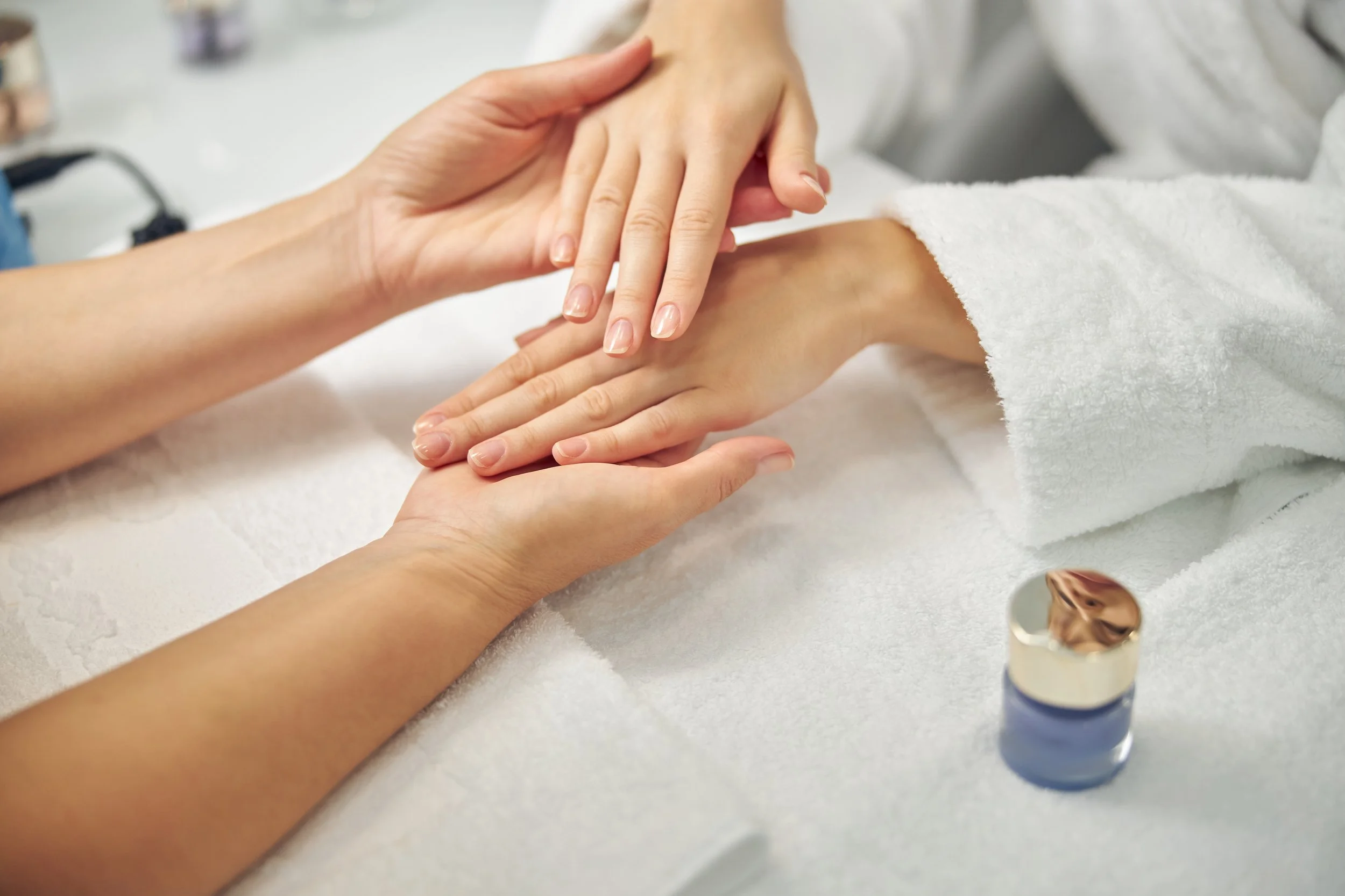 A person receiving a manicure, with a nail technician applying nail polish to their nails. The scene includes hands, a white towel, and a bottle of nail polish on a white surface.