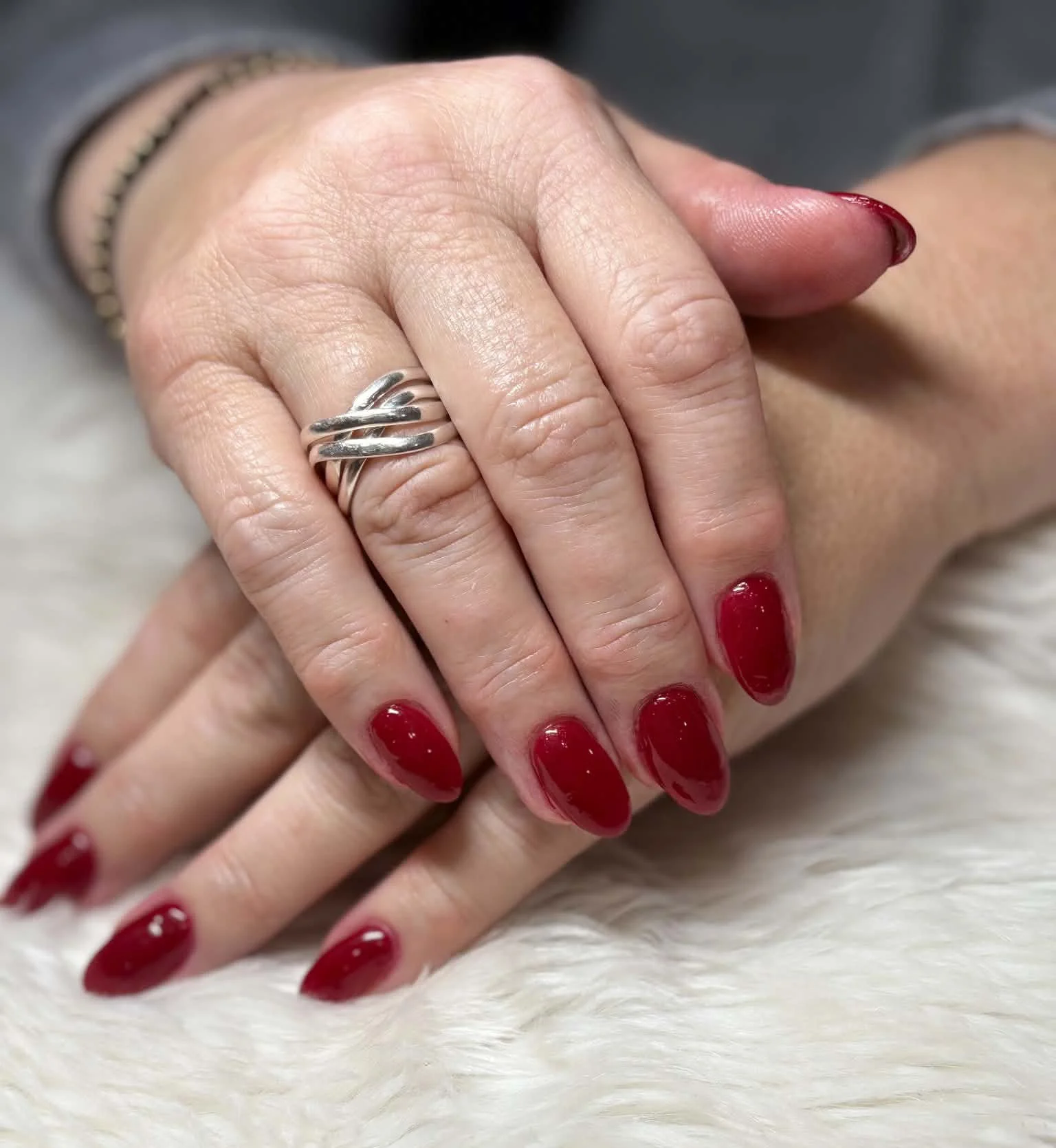 A person's hands with red painted nails, wearing a silver ring with intertwined bands, resting on a soft white surface.