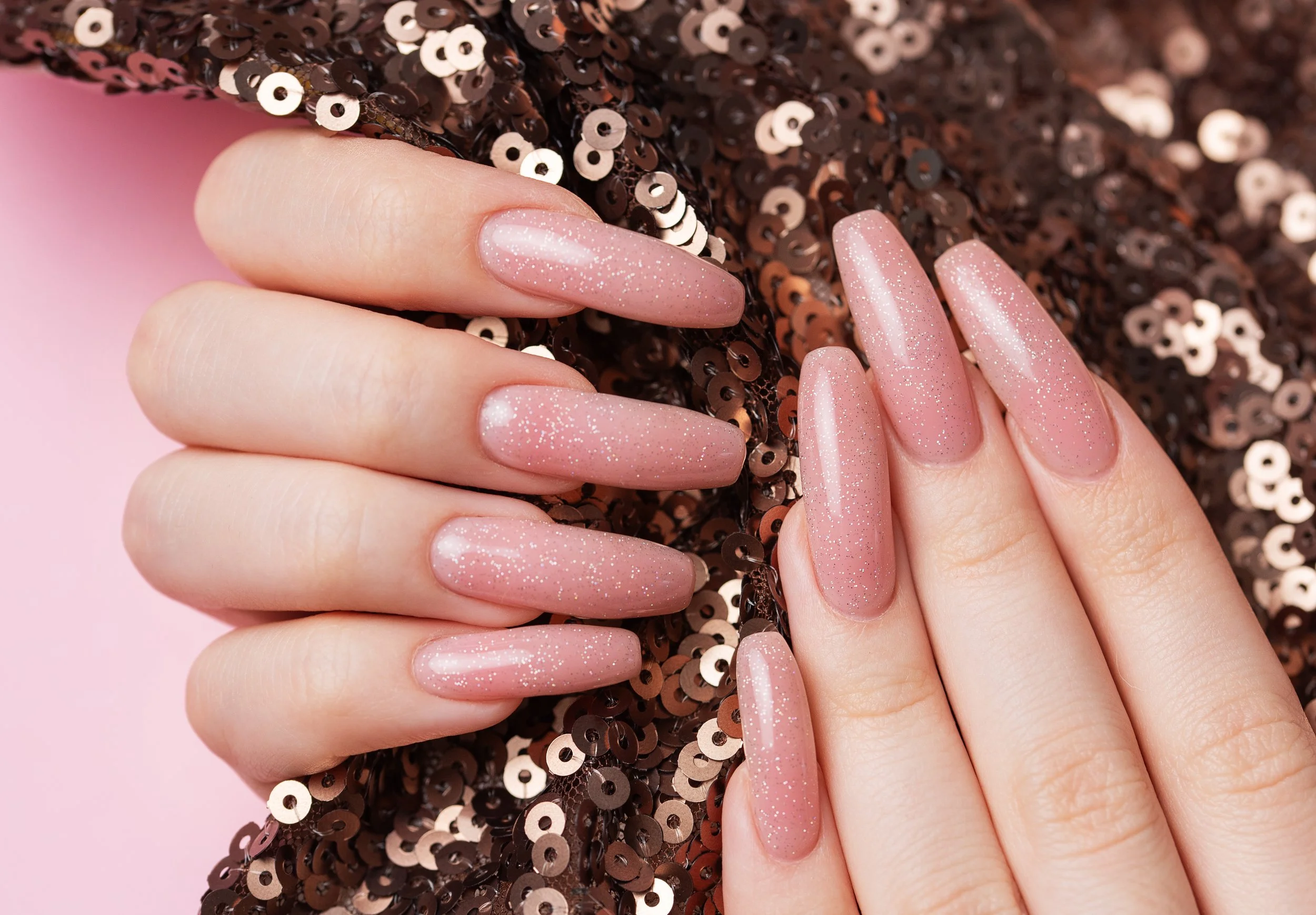 Close-up of a person's hands with long, oval-shaped, pink glittery nail polish nails, resting on a surface decorated with brown sequins.
