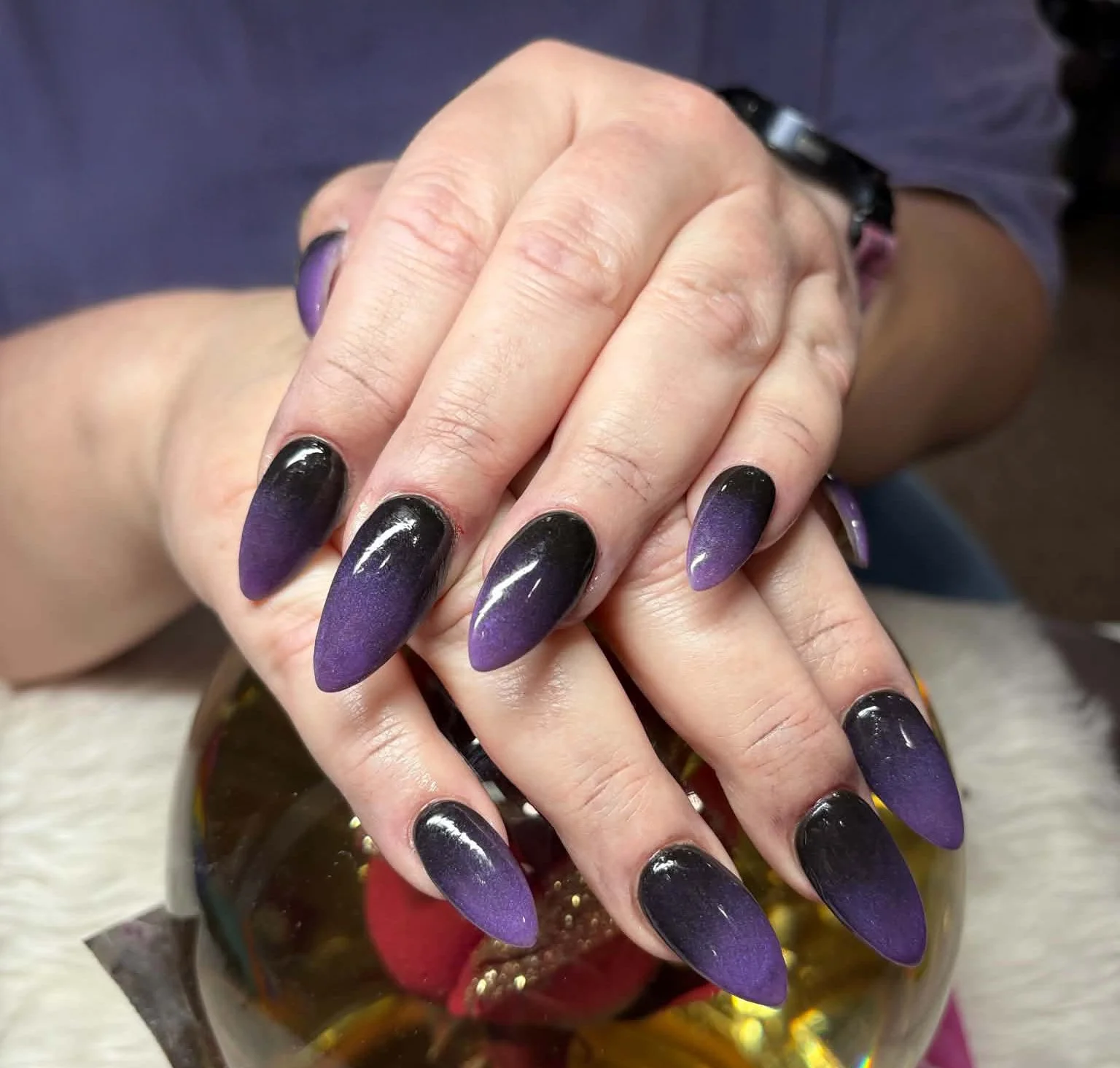 Hands with purple and black ombre stiletto nails resting on a glass orb containing red roses.