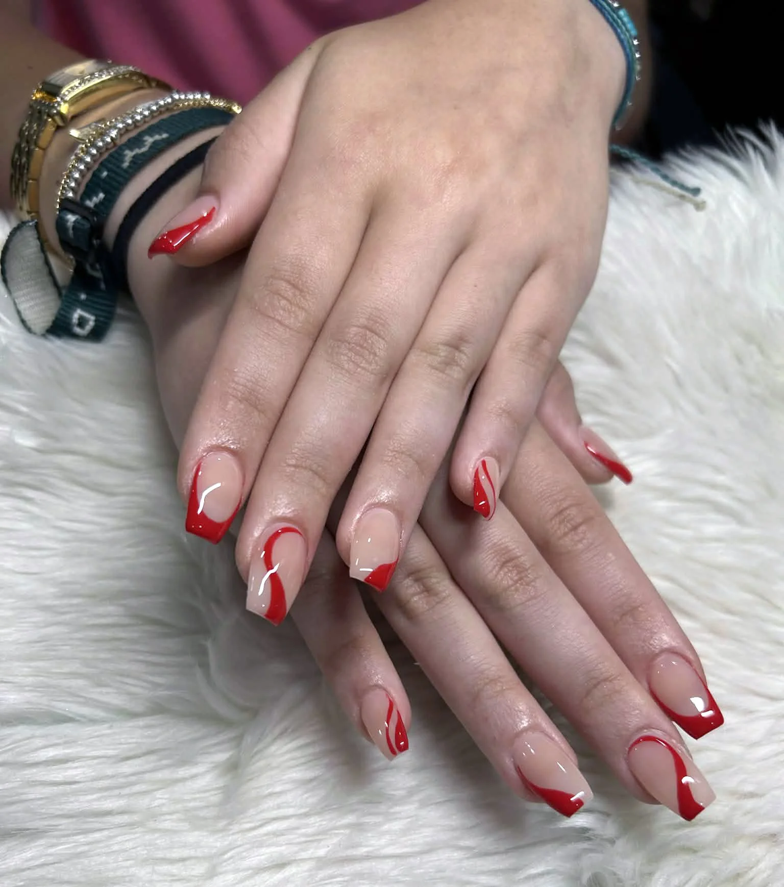 A pair of hands with red and nude color French manicure nails resting on a white furry surface, adorned with multiple bracelets.