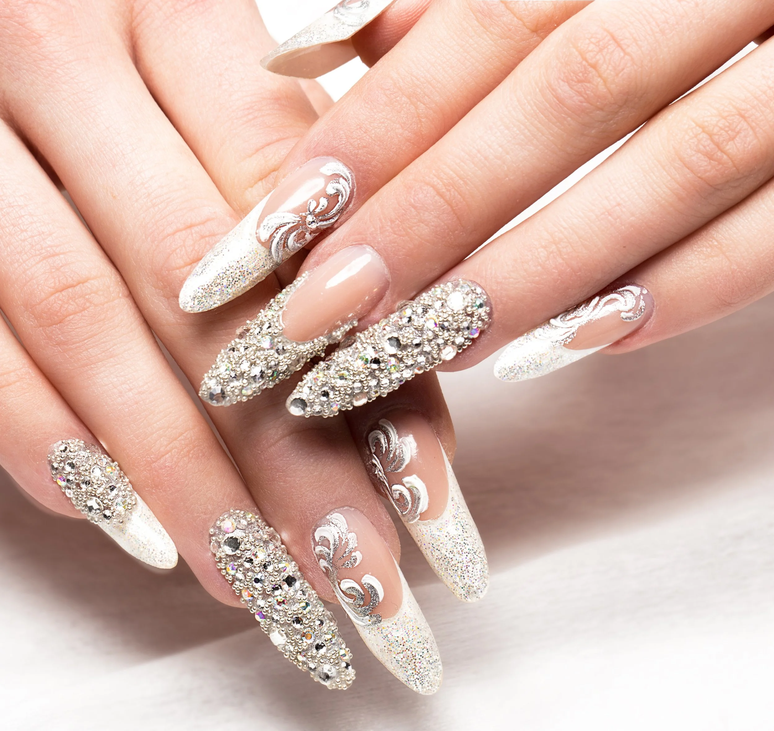 Close-up of hands with long, decorated nails featuring rhinestones, glitter, and intricate white and silver designs.