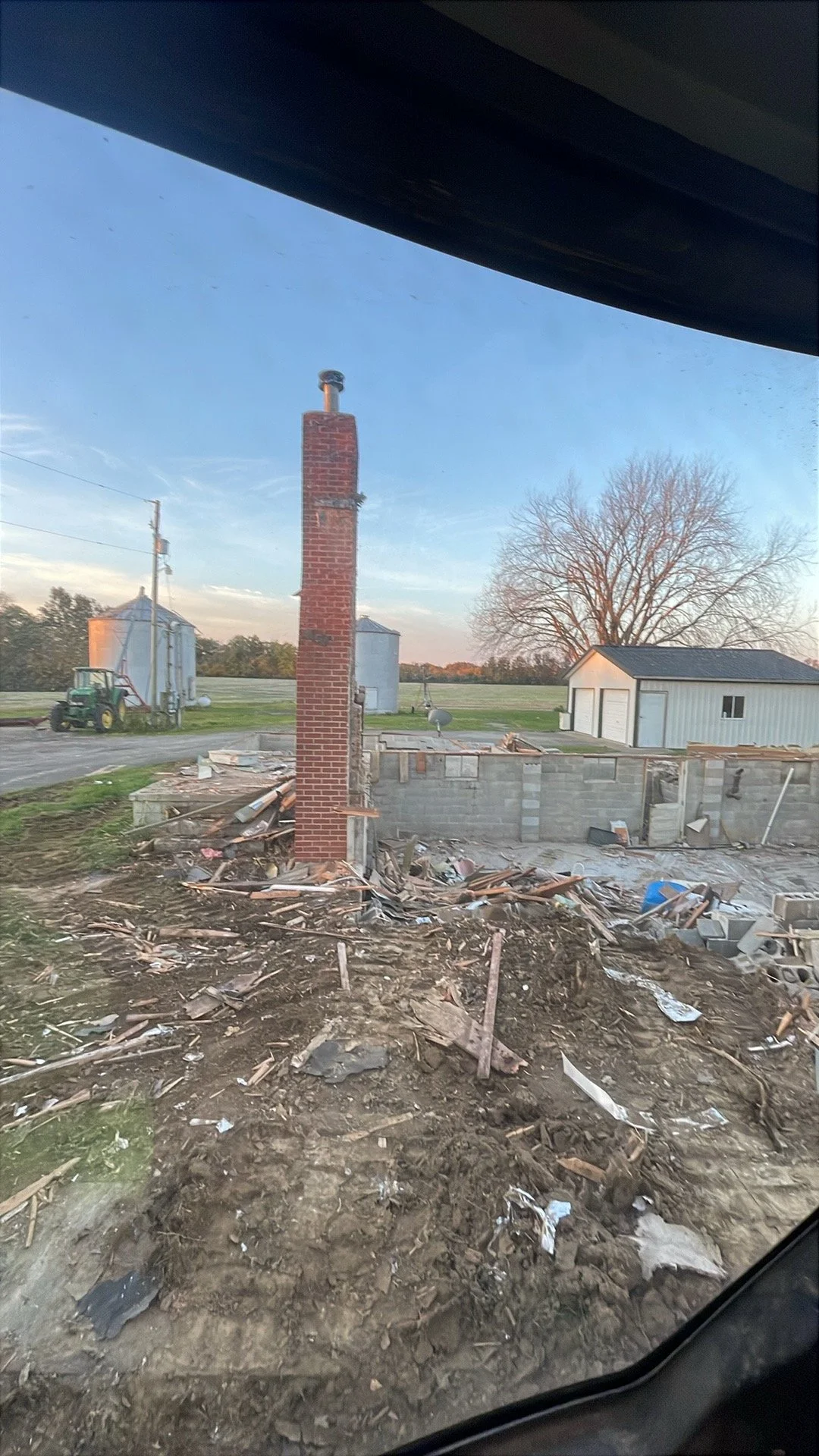 View of a construction site with a brick chimney, scattered wood debris, a partially built wall, and some farm buildings in the background during daylight hours.
