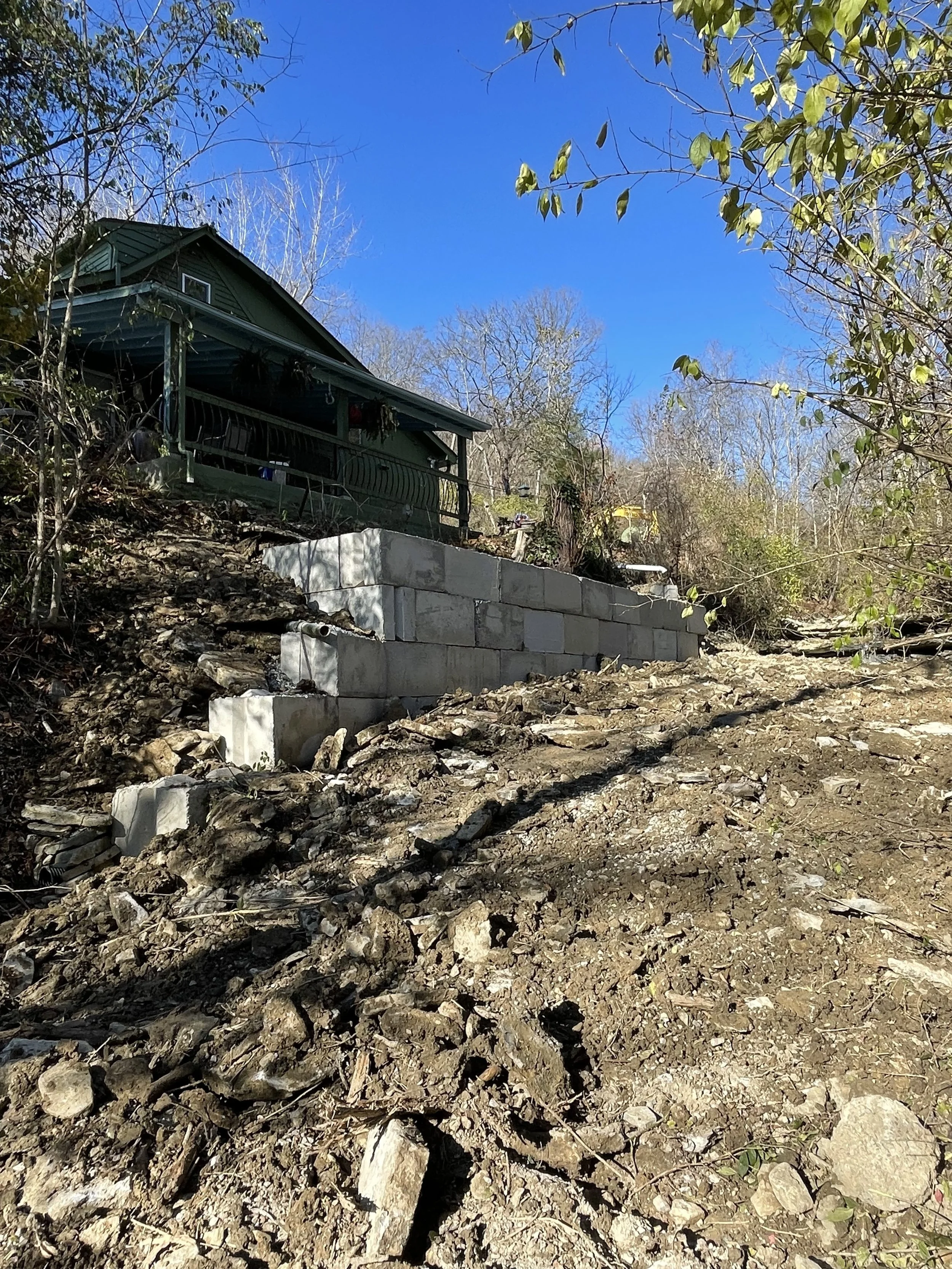 A house on a hill with a green exterior and a porch, with a retaining wall made of concrete blocks in front, surrounded by trees with bare branches and some green leaves, under a clear blue sky.