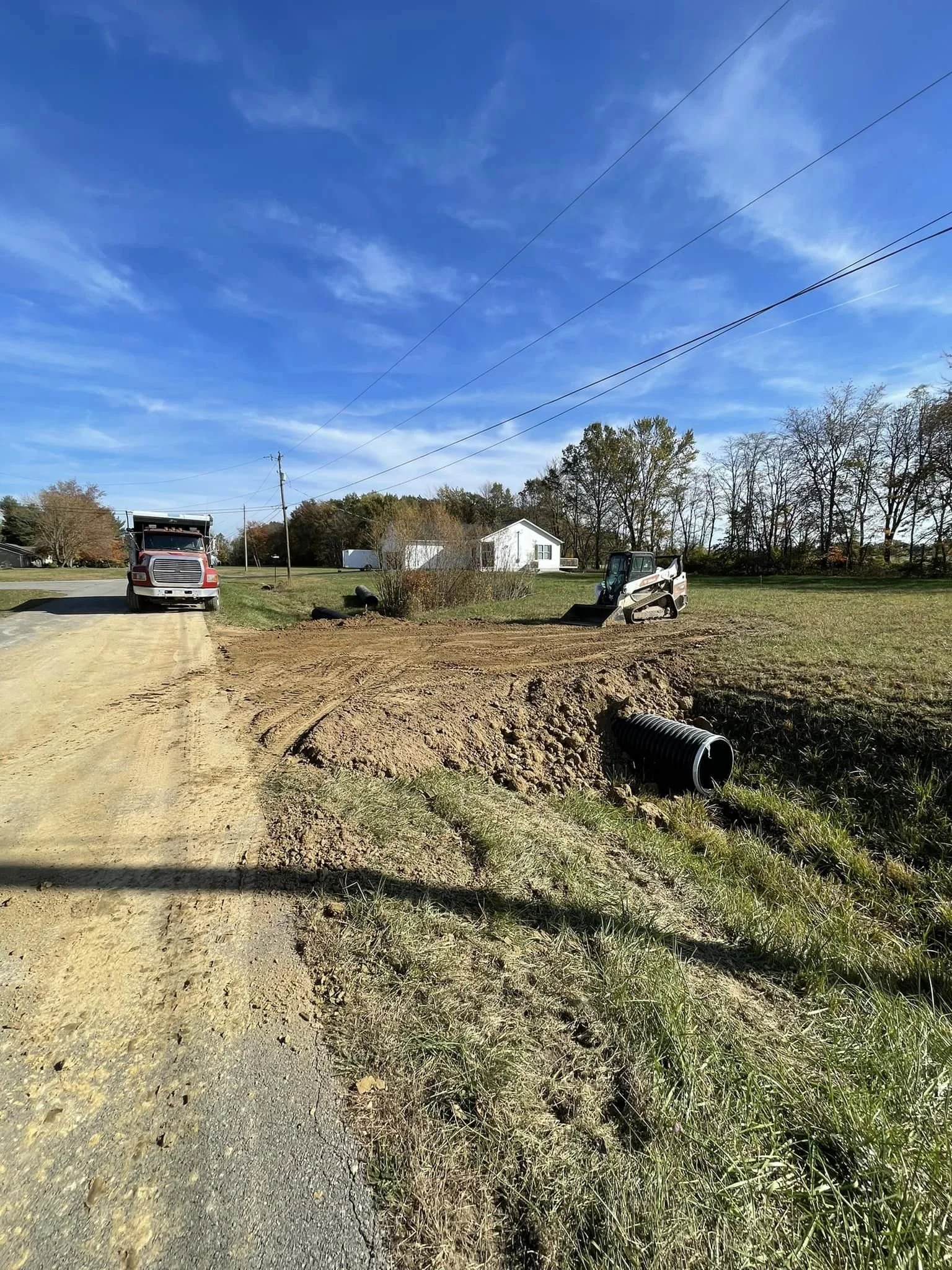 Construction site on a rural road with a small bulldozer, a truck, and a large drainage pipe.
