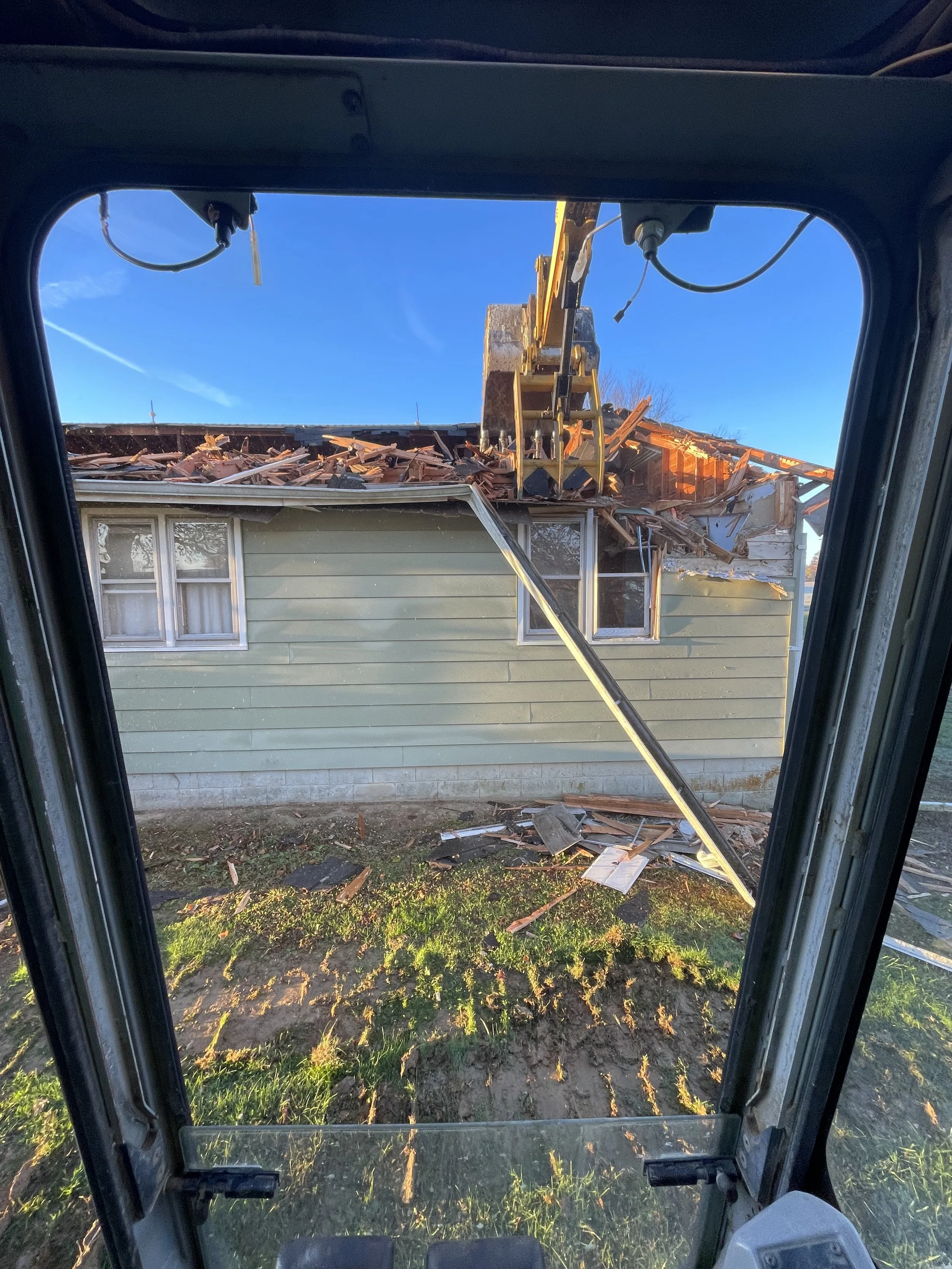 Construction excavator demolishing a house, viewed from inside the cab with the door open. The house has light green siding, two windows with white trim, and parts of the roof are being torn off. Debris from the roof is scattered on the ground.