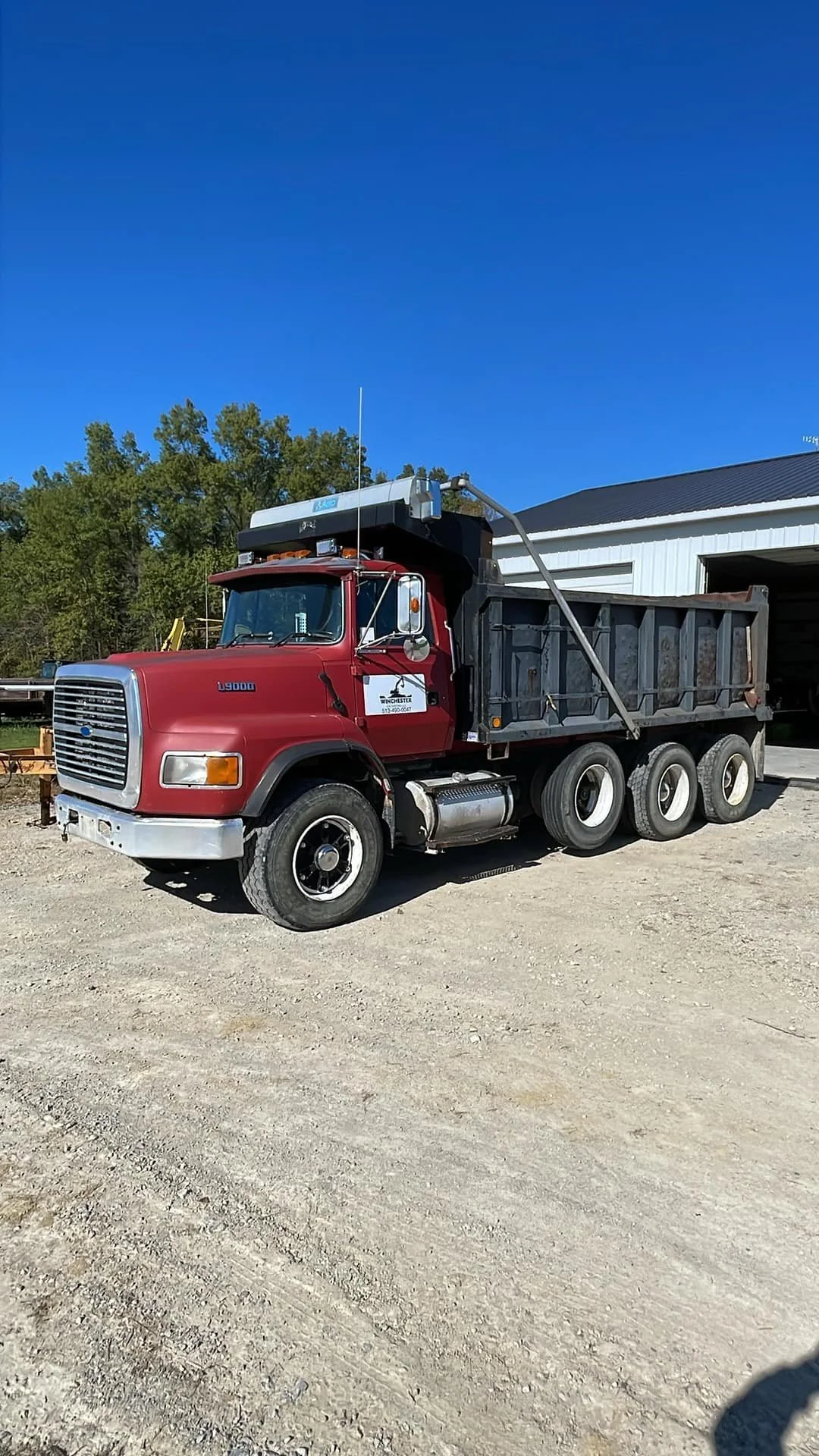 Red dump truck parked on dirt ground near a white building with trees in the background under a clear blue sky.