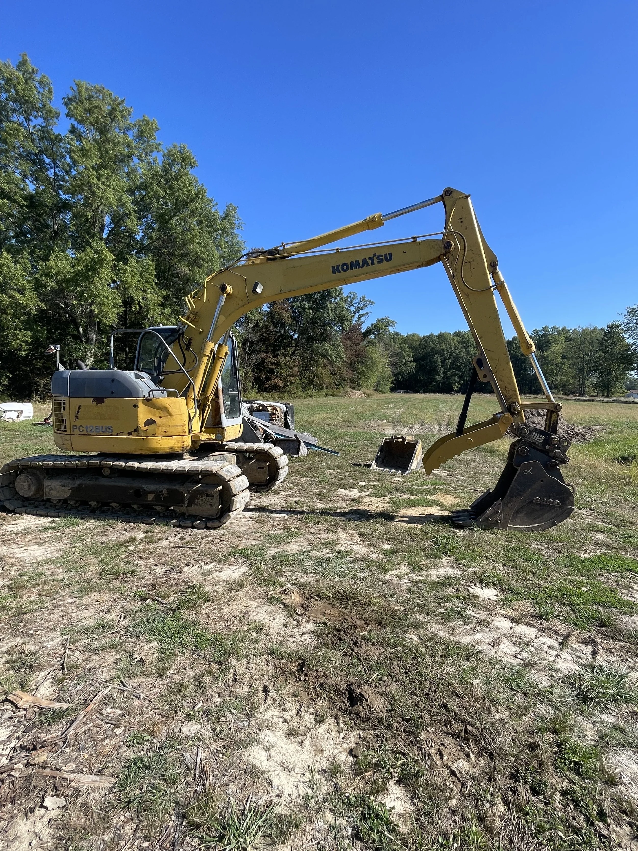 Yellow Komatsu excavator with black bucket attachment resting on dirt and grass in an open field, with trees and a clear blue sky in the background.