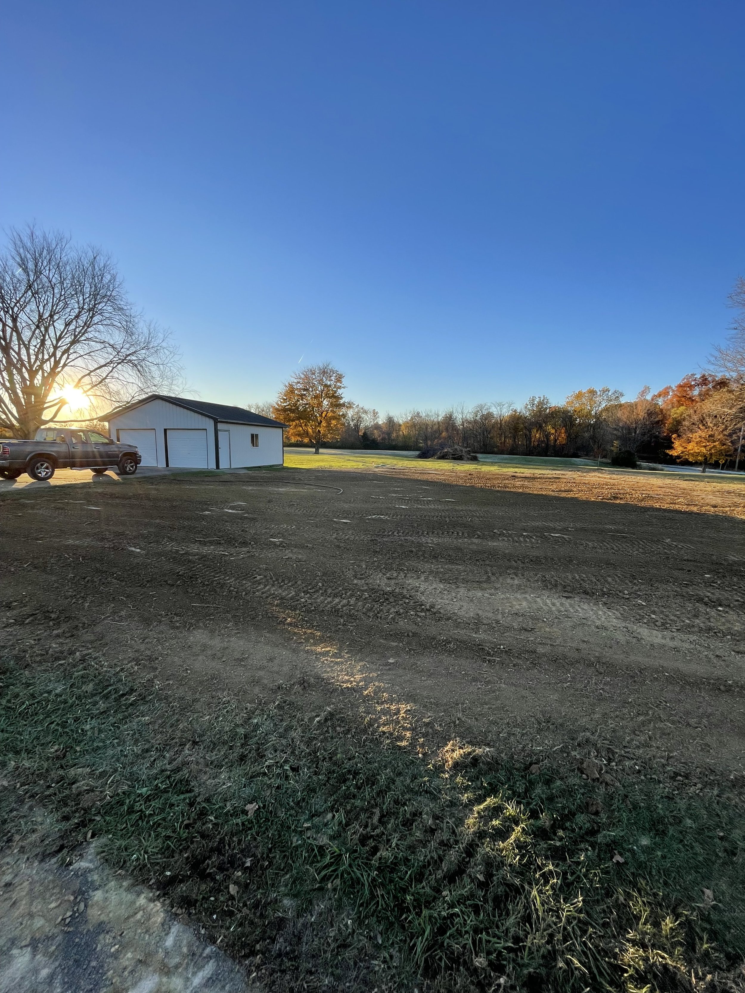 Open dirt lot with a white building and two parked trucks, surrounded by trees with autumn leaves, under a clear blue sky at sunset.