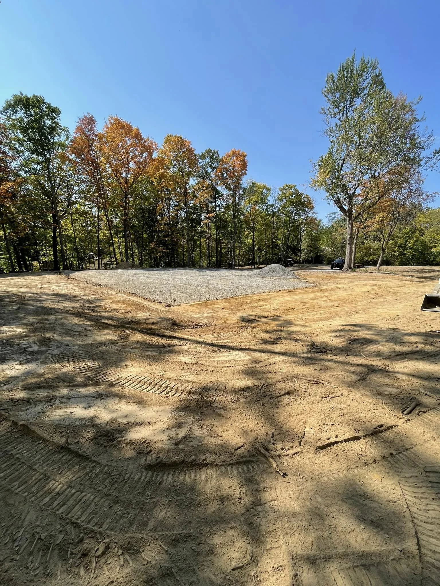 A cleared and leveled patch of ground with gravel in the center surrounded by bare dirt, with trees in the background and a clear blue sky.
