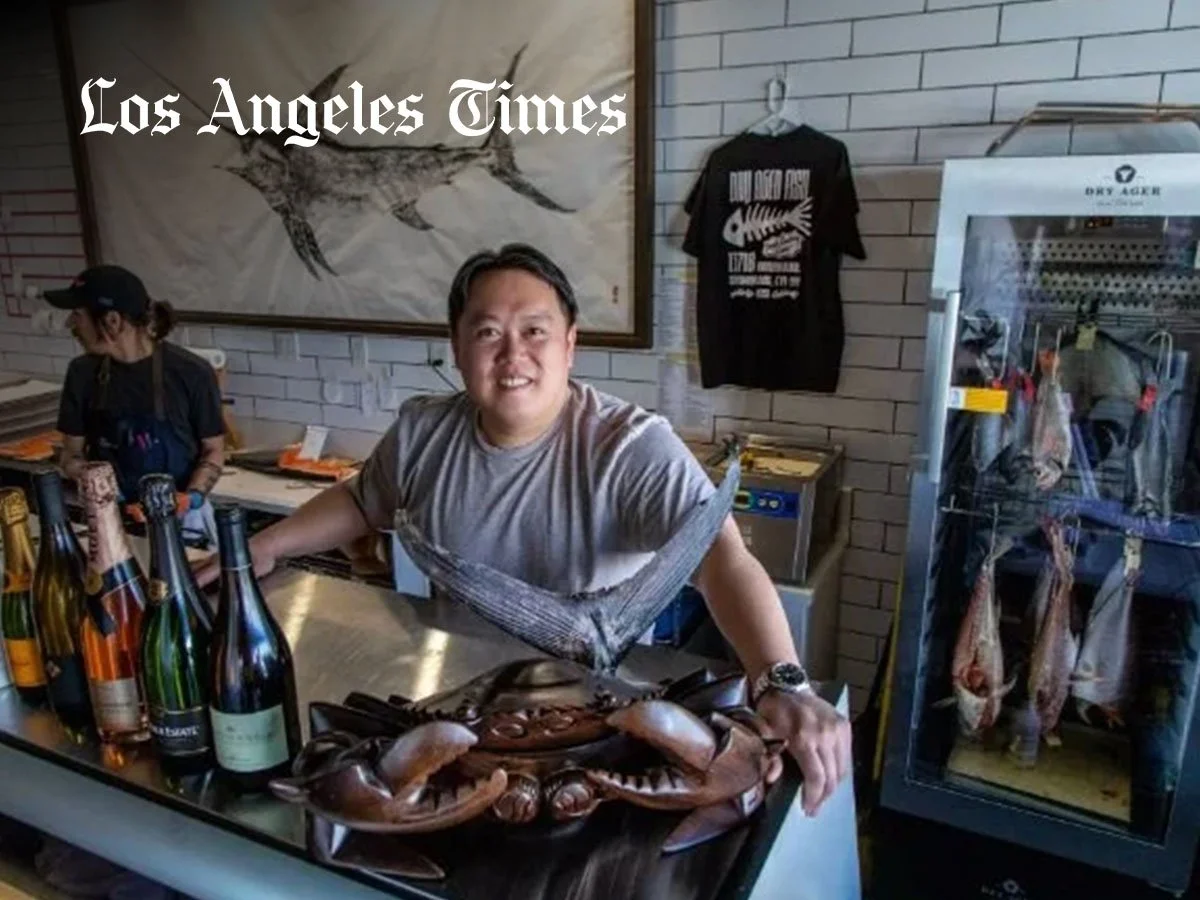 A cheerful man at a seafood restaurant counter displays a large crab. Behind him, a woman works, and on the wall is a fish illustration. To the right, a refrigerated display case holds hanging fish and seafood.