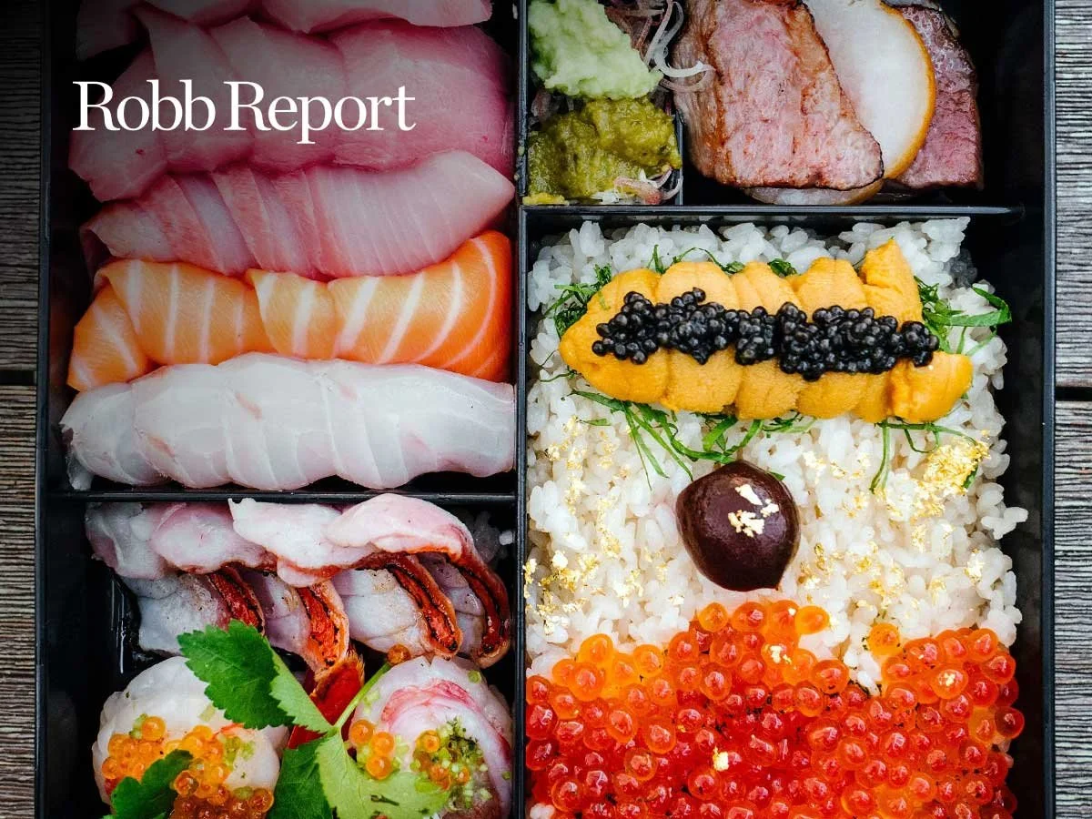A Japanese bento box with assorted sashimi, rice topped with sea urchin, caviar, and salmon roe, served in a black compartmentalized container.