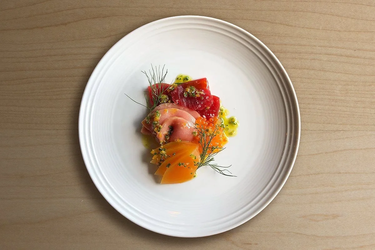A white plate with slices of colorful raw fish or sashimi, garnished with herbs and small green and yellow accents, on a wooden surface.