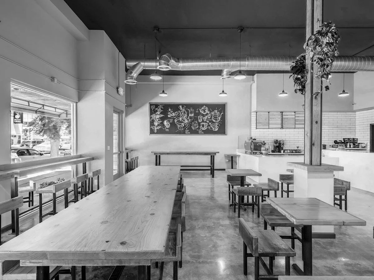 Interior of a modern coffee shop with wooden tables and chairs, a chalkboard with illustrations, and a counter area with coffee equipment.