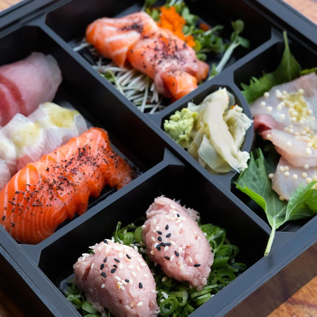 A black bento box with six compartments containing slices of raw salmon, white fish, tuna, garnished with black and white sesame seeds, and accompanied by vegetables including shredded cabbage, leafy greens, and microgreens.