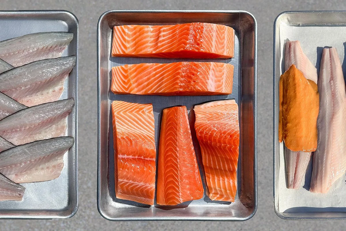 Trays of fresh fish fillets, including salmon, arranged on metal trays.