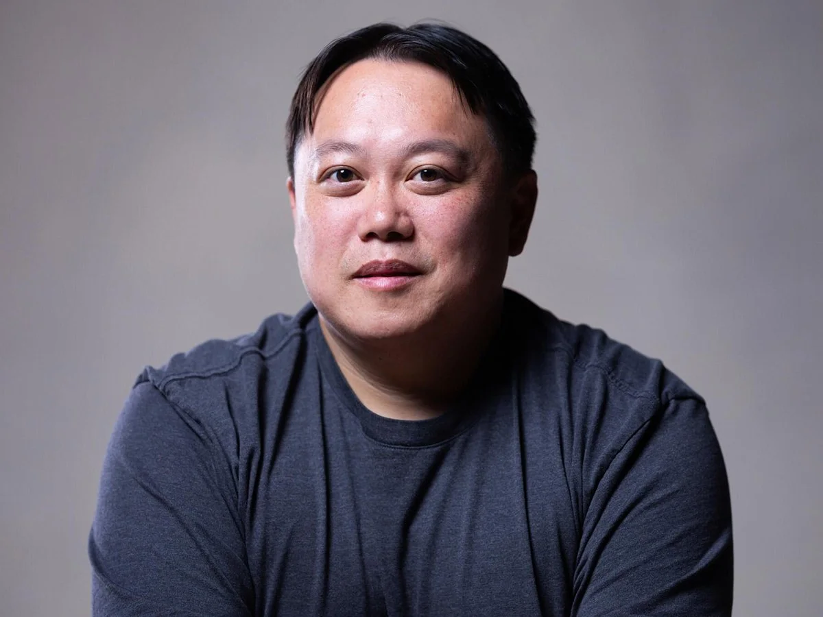 Portrait of a man with short black hair, wearing a dark gray t-shirt, looking directly at the camera against a plain background.