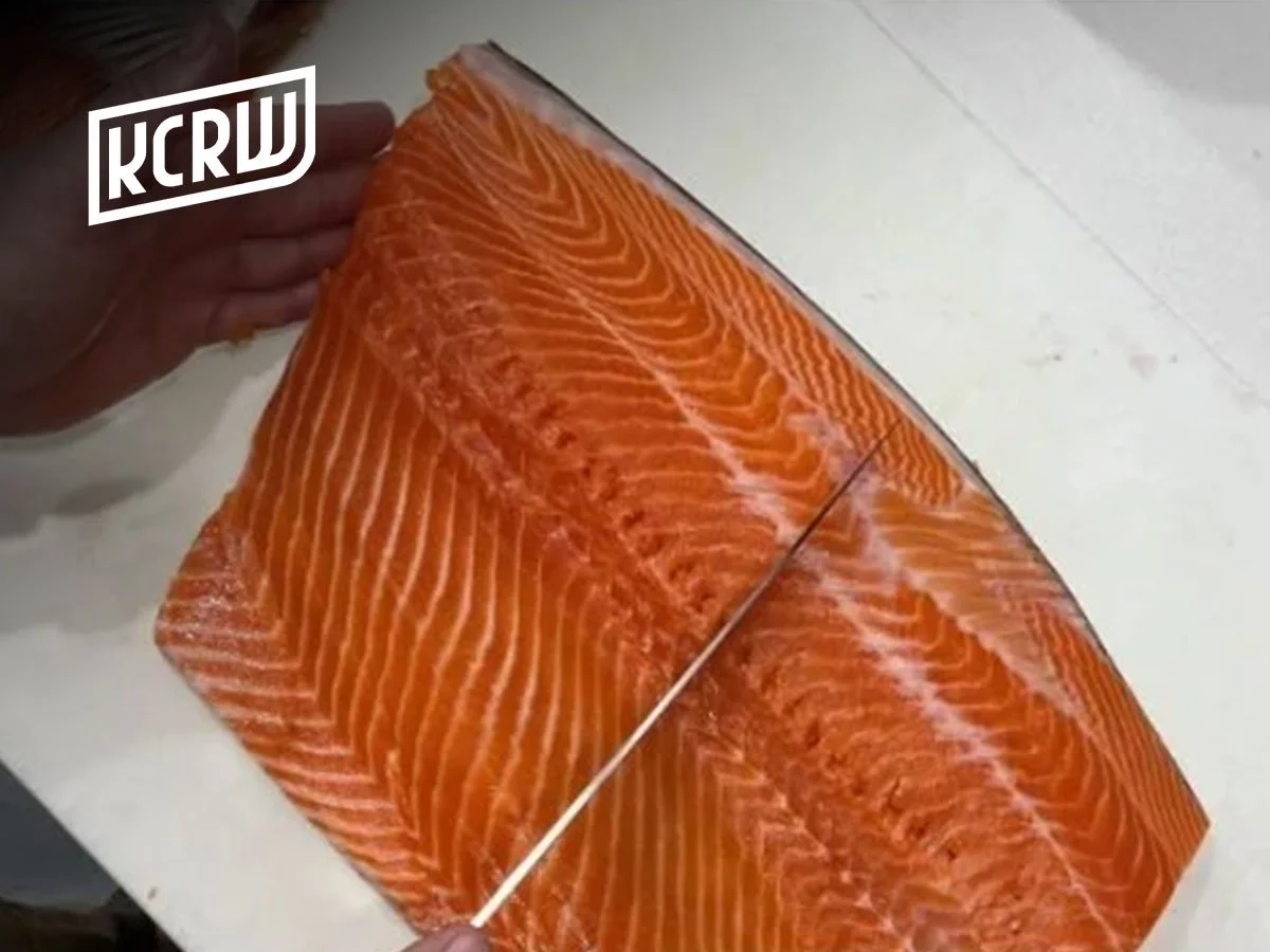 A person cutting a large fillet of raw salmon with a knife on a white surface.