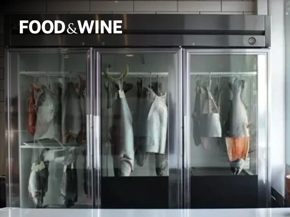 A commercial refrigerator with glass doors containing hanging cuts of various meats, labeled as food and wine.