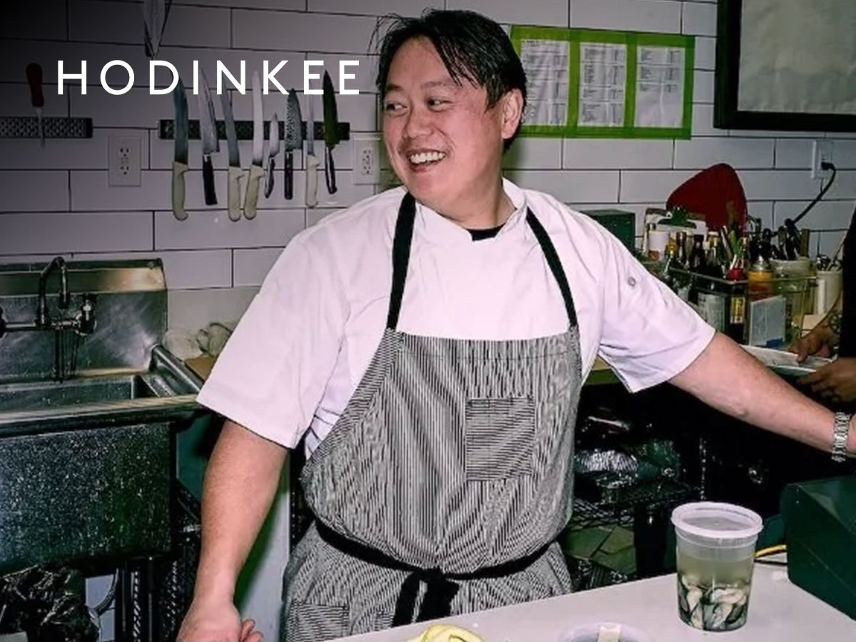 A smiling chef wearing a striped apron in a commercial kitchen, with knives hanging on the wall behind him and various kitchen tools and ingredients on the counter.