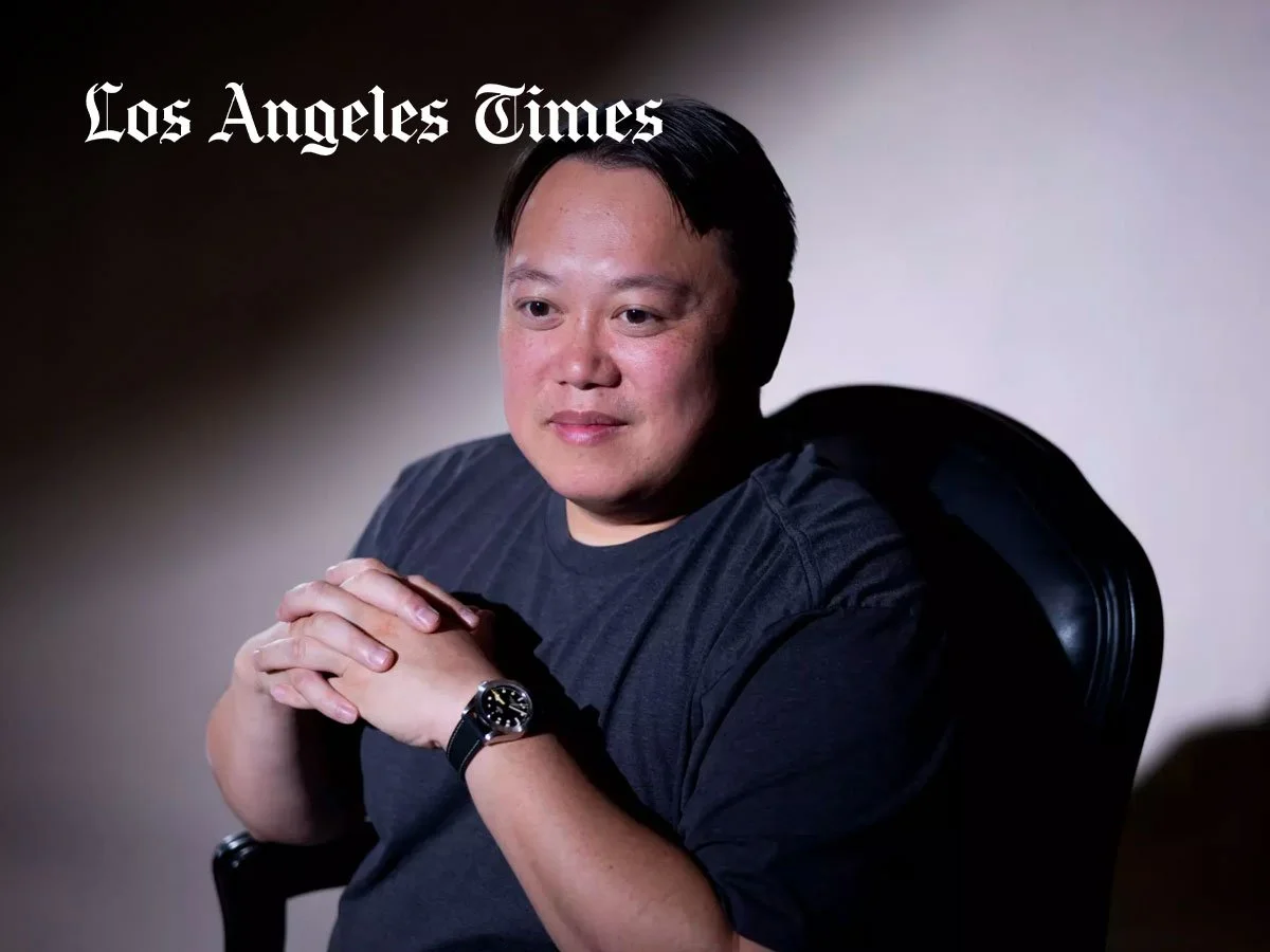 A man sitting in a chair with his hands clasped, wearing a black shirt and a watch, with a gradient background. The logo 'Los Angeles Times' is in the top left corner.