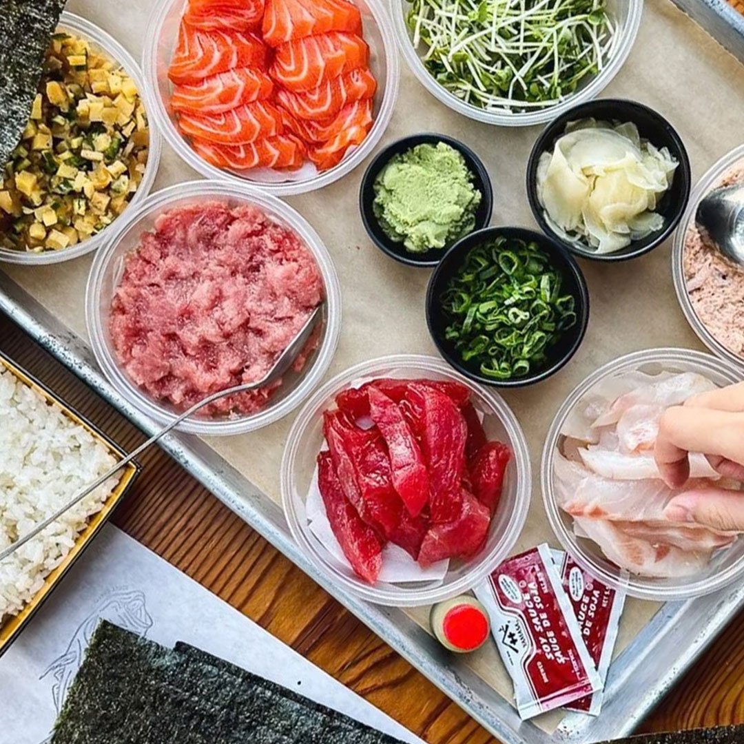 Assorted Japanese sushi ingredients including sliced salmon, tuna, raw fish, avocado, chopped green onions, shredded cabbage, pickled ginger, wasabi, microgreens, and sushi rice, on a tray.