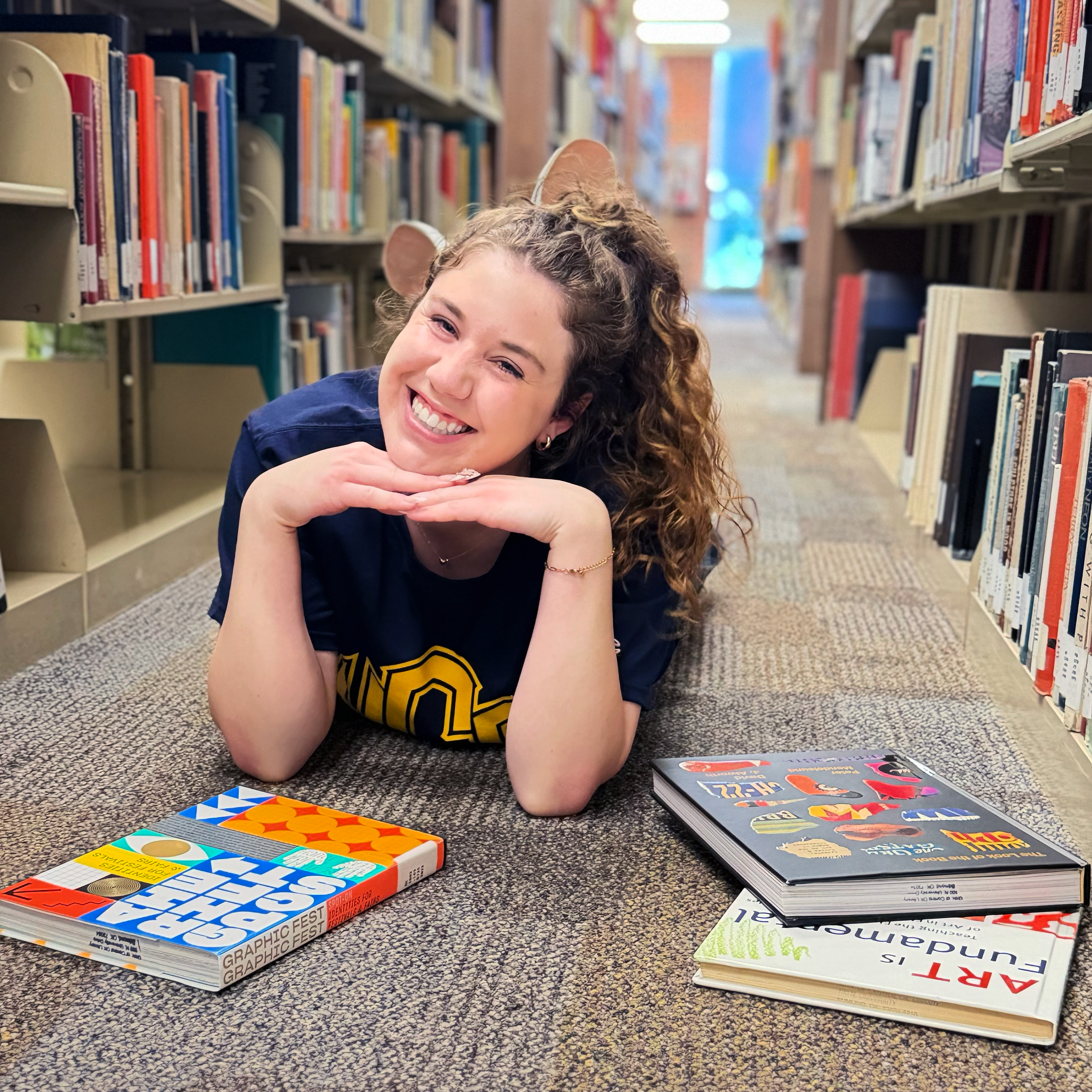 A smiling girl with curly hair and animal ears laying on the floor, surrounded by books on shelves in a library.