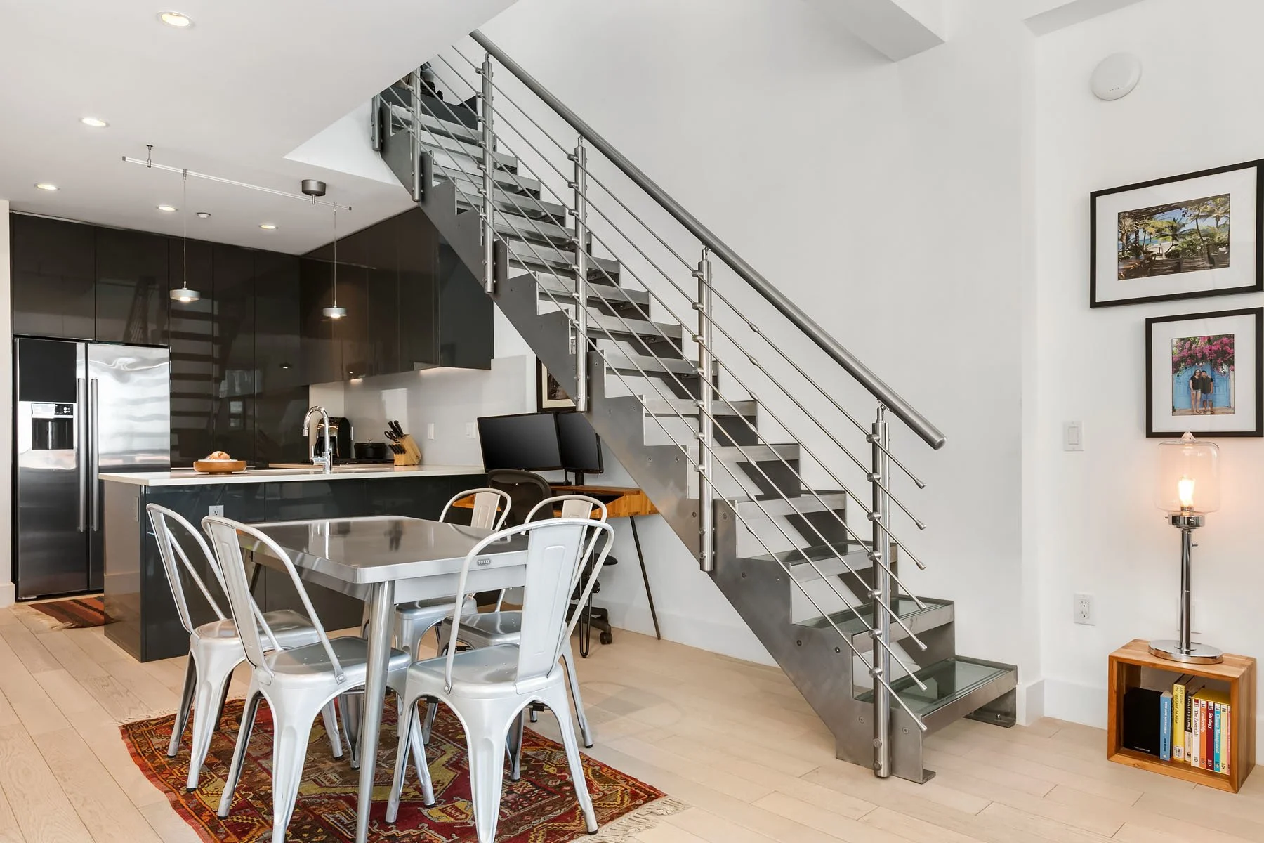 Modern kitchen with black cabinets, stainless steel appliances, breakfast bar, and dining table, with a metal staircase leading to the upper floor and framed photos on a white wall.