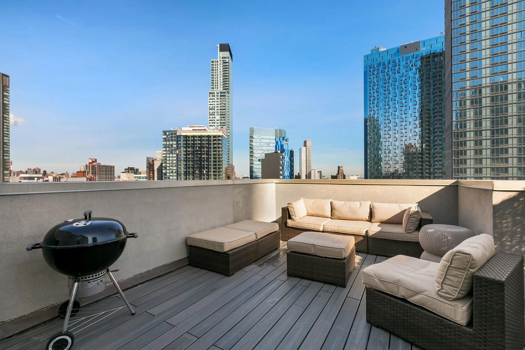 Rooftop patio with outdoor furniture and a barbecue grill, overlooking a city skyline with tall modern buildings under a clear blue sky.