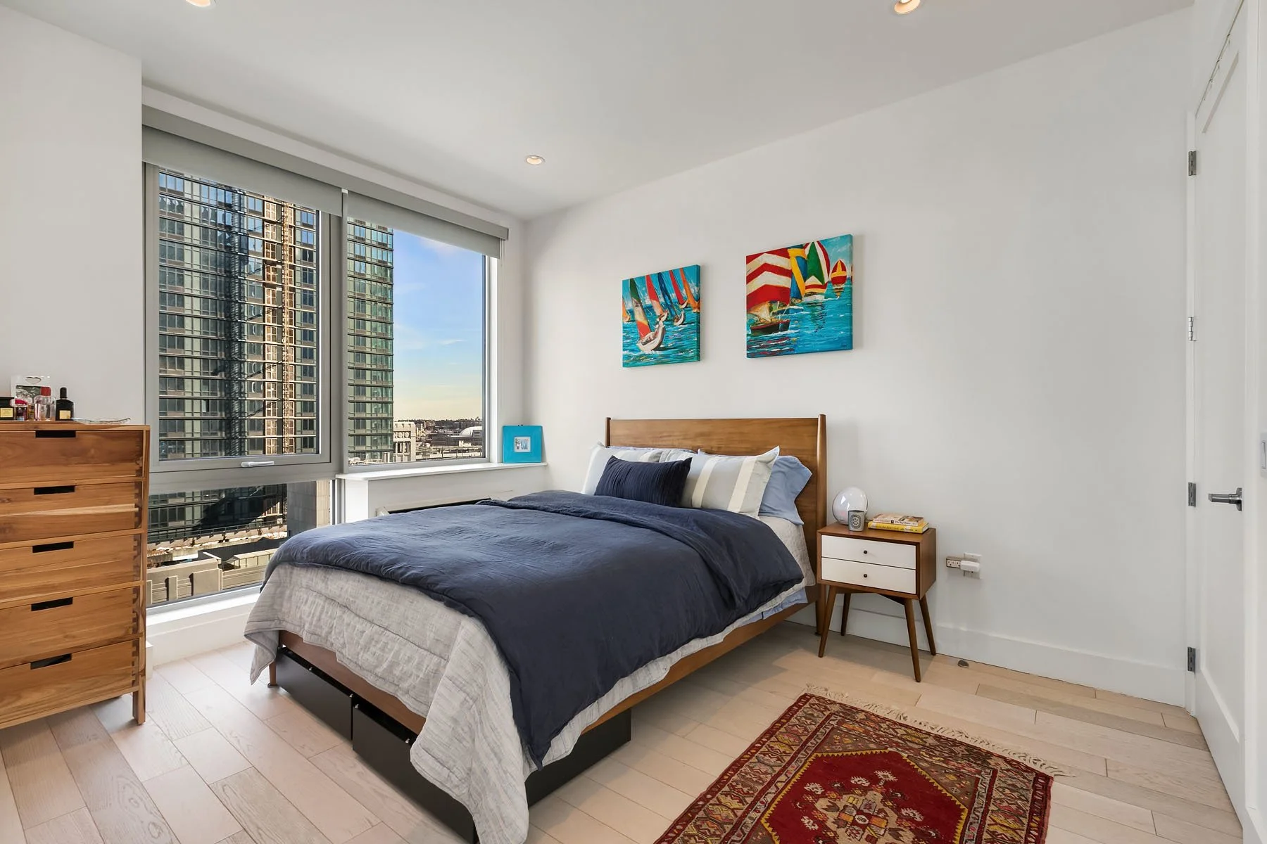 Modern bedroom with a large window showing city high-rise buildings, a bed with navy blue and beige bedding, artwork of sailboats on the wall, a wooden dresser, and a nightstand with books and a lamp.
