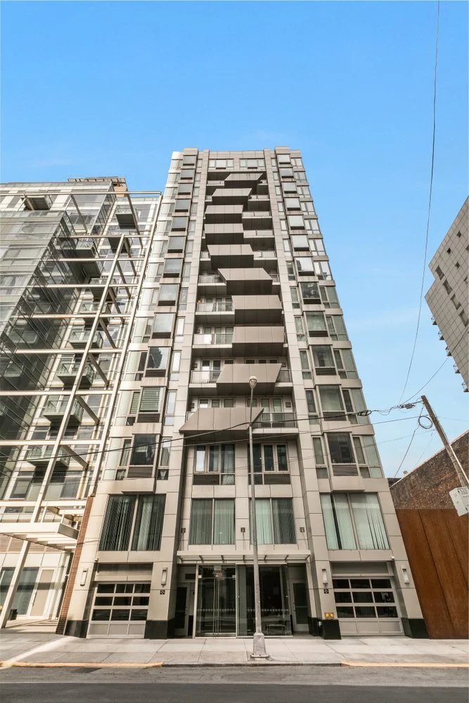 Tall modern residential building with balconies, adjacent to other multi-story buildings, under a clear blue sky.