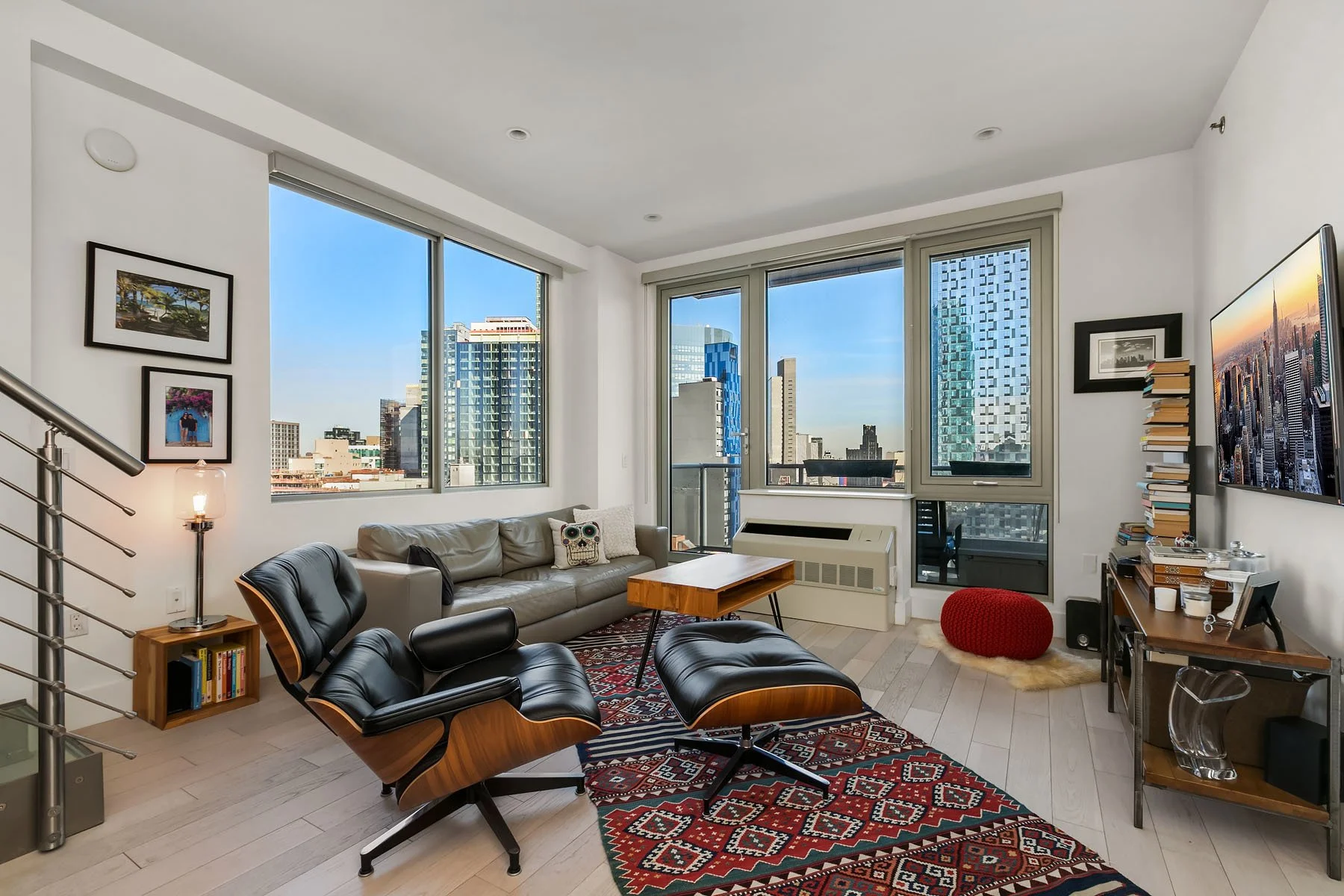 Living room with large cityscape view through tall windows, featuring modern furniture including black lounge chairs, a gray sofa with a decorative skull pillow, a wooden coffee table, and a colorful patterned rug. There are framed photographs on the walls, a red pouf on a sheepskin rug, and a television mounted on the wall. A small bookshelf and a side table with a lamp are also visible.