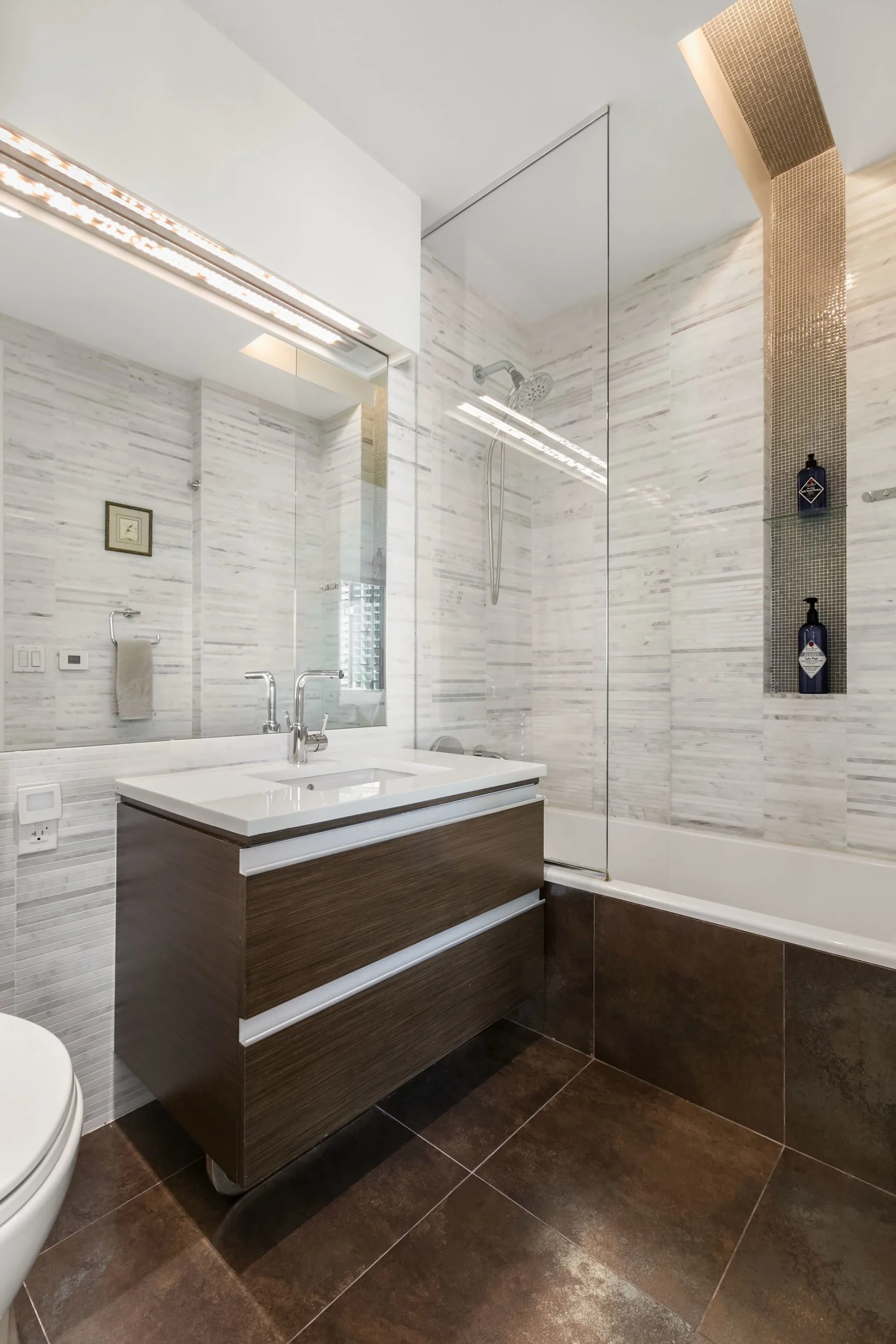 Modern bathroom with white marble walls, a large mirror, a white sink with a wooden vanity, a bathtub with a glass shower partition, and built-in shelves with bottles of shampoo.