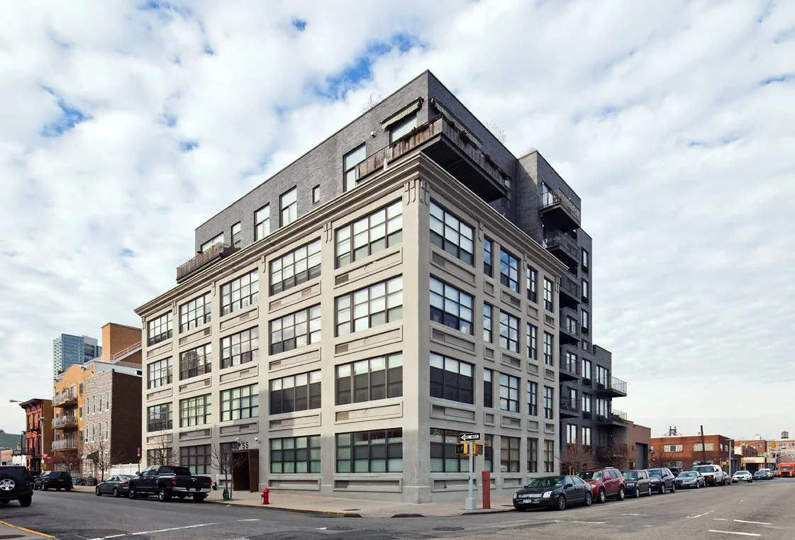 A multi-story urban building with large windows, some balconies, situated on a street corner with parked cars and a cloudy sky.