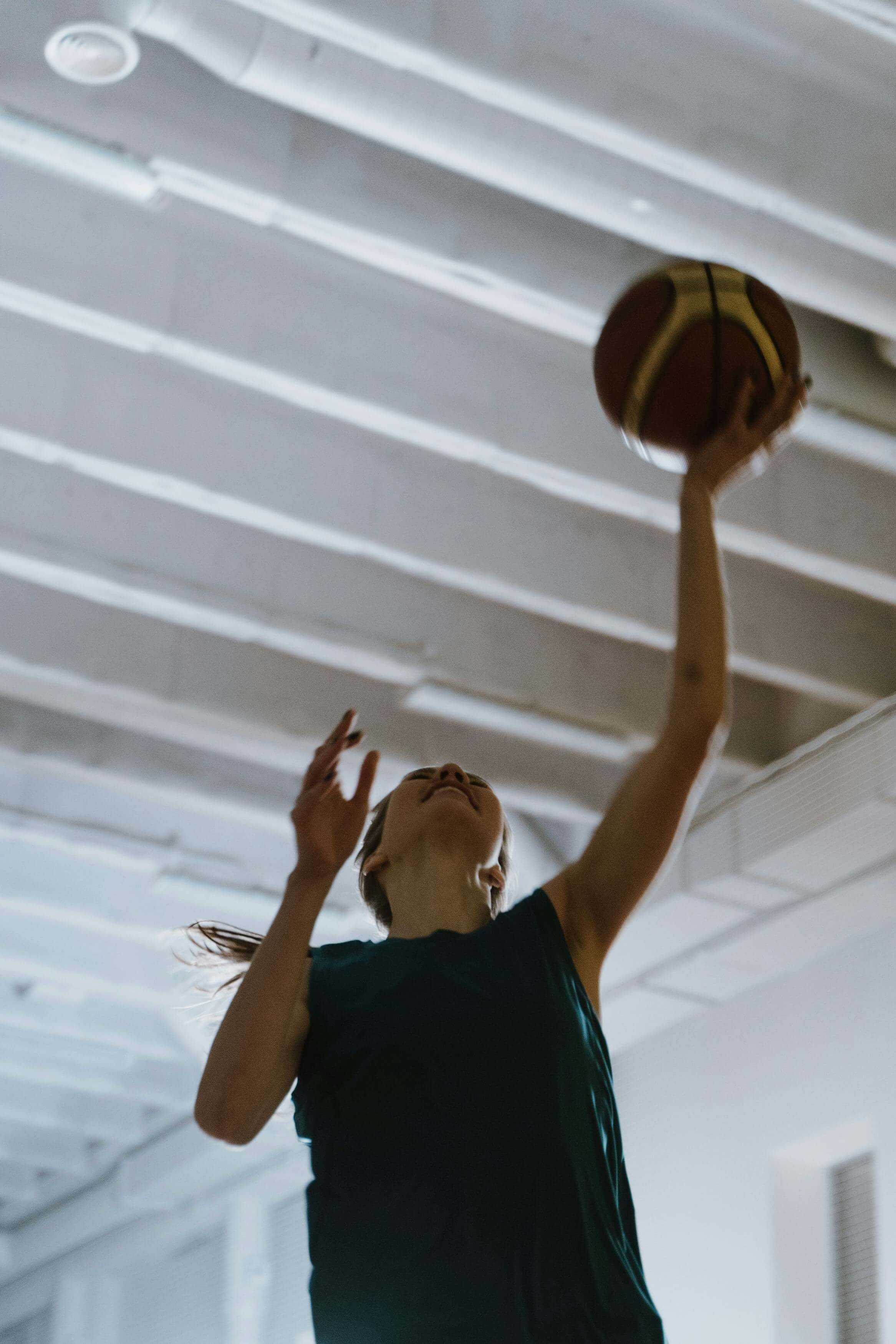 A woman playing basketball indoors, reaching up with one arm to shoot or dunk the ball.