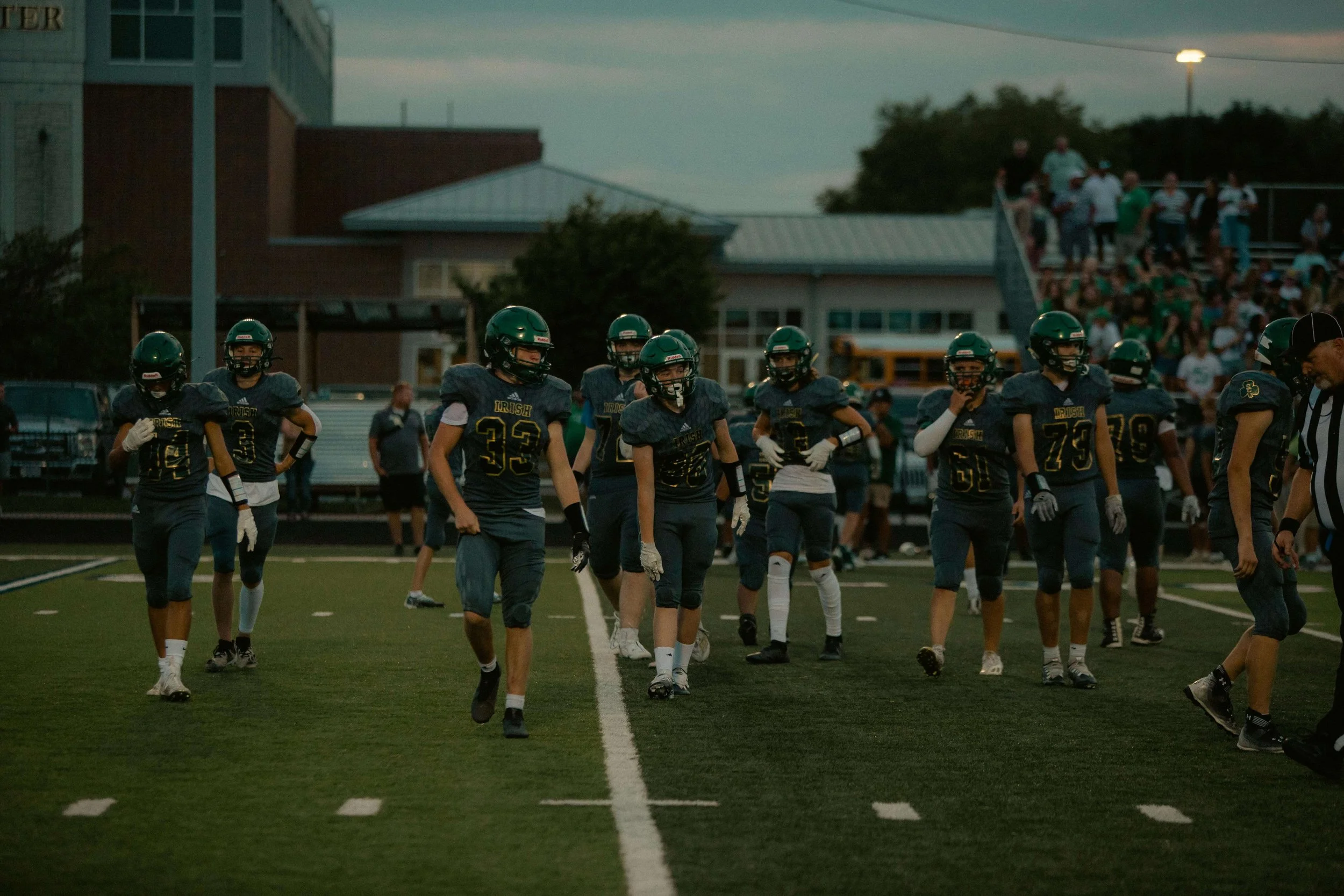 Young football players in dark uniforms and helmets standing on a football field during a game with spectators in the bleachers and buildings in the background.